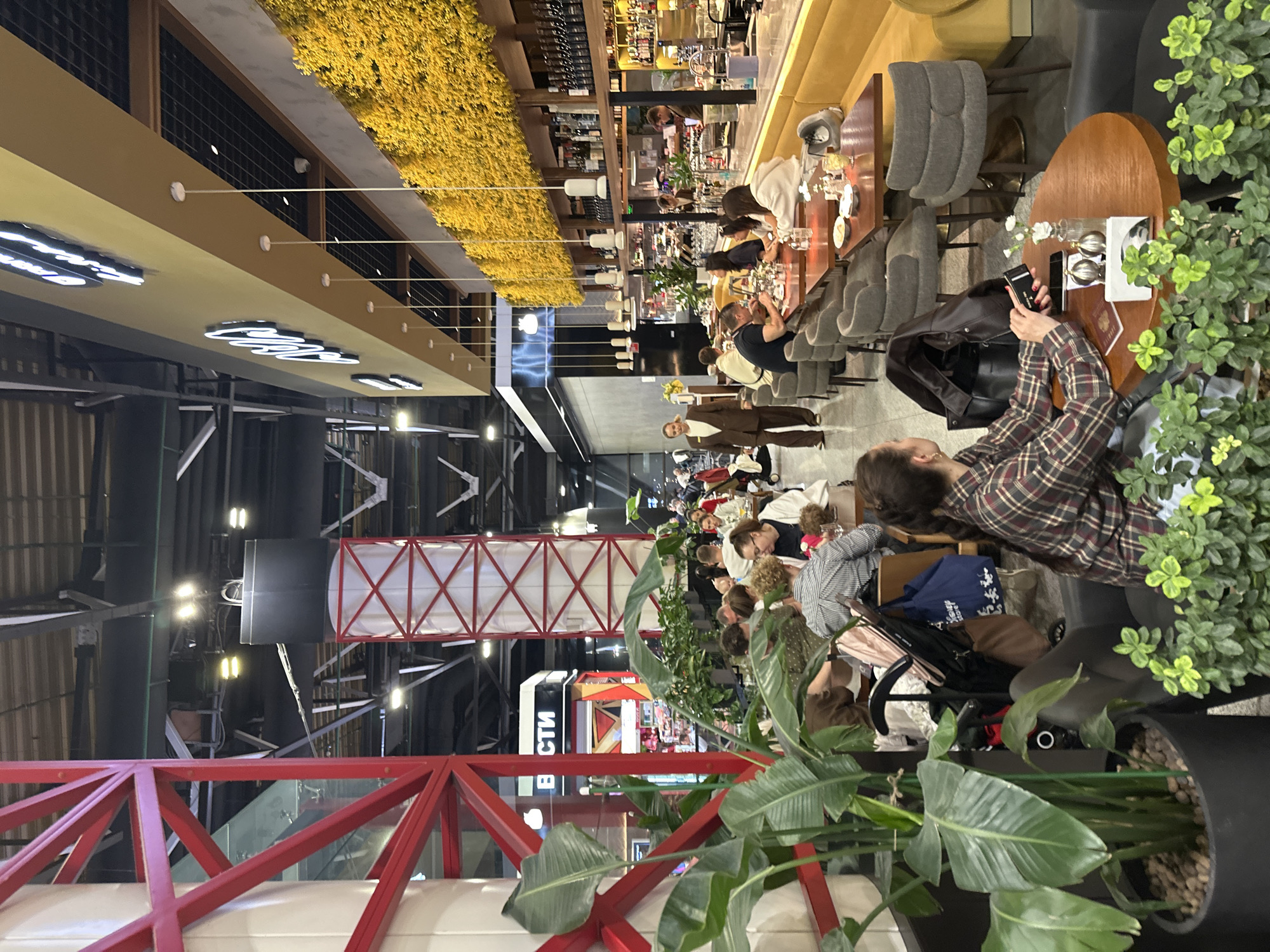 A horizontal indoor shot reveals a bustling scene in what appears to be a restaurant or dining area within a larger venue, potentially in Khimki, Russia. The image is oriented with the right side facing the camera.
In the foreground, large potted plants with lush green foliage partially obscure the view, framing the central activity. A person dressed in a plaid shirt is seated at a wooden table and appears to be taking a picture with their smartphone. The table is also adorned with decorative plants, glasses, and other tableware. A brown leather jacket is draped over the chair.
The restaurant is filled with patrons, seated at various tables. There are diverse groups of people, including individuals, couples, and larger parties. The seating arrangements are varied, with some tables featuring plush, upholstered chairs and others with simpler designs.
Overhead, a long bar area is visible, stocked with bottles of alcohol and other beverages. Above the bar, there is an extensive yellow floral arrangement. The ceiling of the building reveals an industrial aesthetic with exposed beams, red metal architectural support structures, and various fixtures.
The environment suggests a modern, perhaps upscale dining establishment within a larger commercial or public space. The image captures the lively atmosphere of a gathering place.