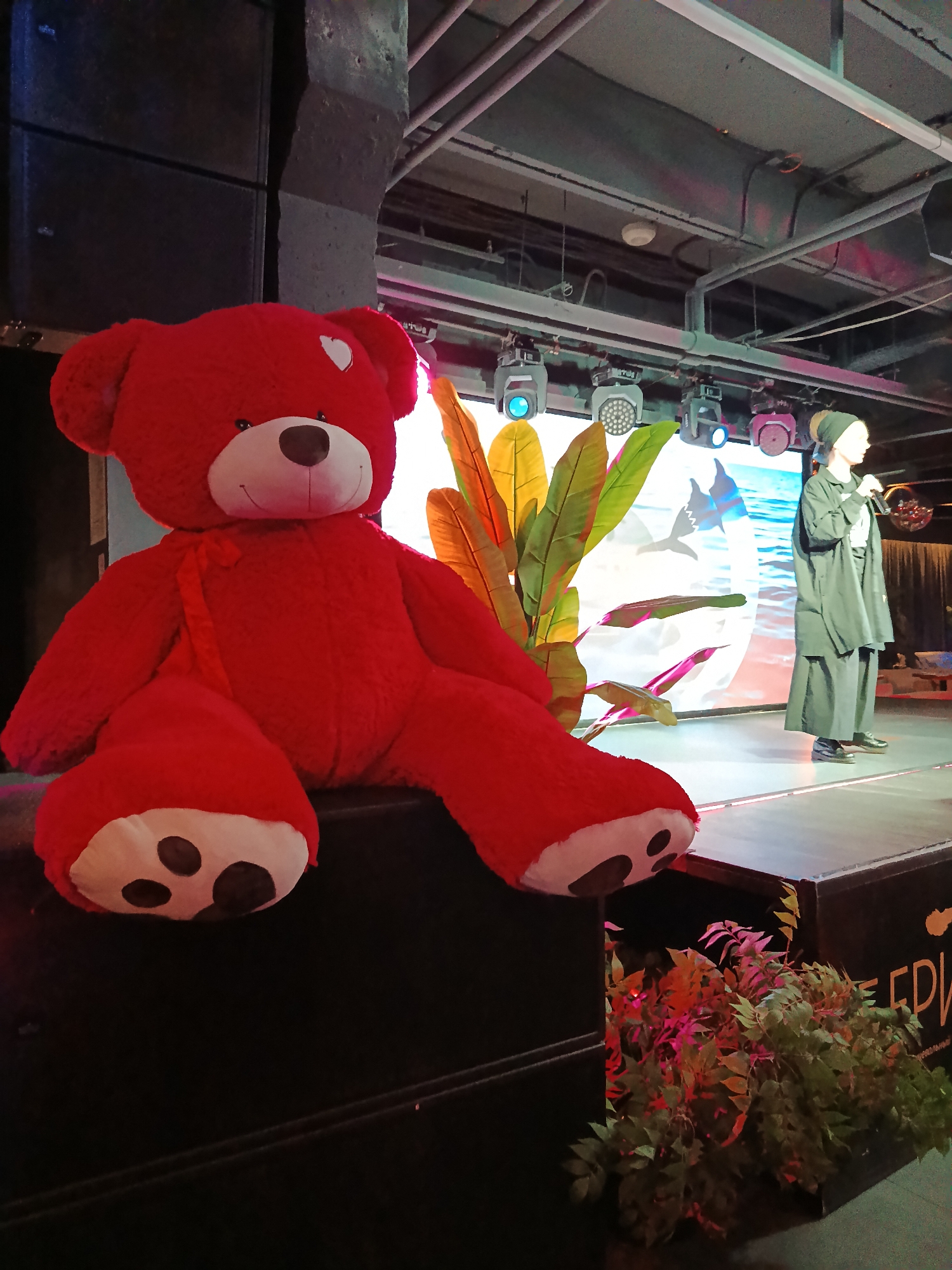 A brightly lit interior scene features a large red teddy bear prominently placed in the foreground, perched on top of a large black speaker. The teddy bear has white paw pads with dark brown markings and a white heart on its right ear. In the background, a woman wearing a dark green headscarf and a dark green oversized coat and wide-legged pants stands on a brightly lit stage, holding a microphone. Behind her, a screen displays a graphic design of ocean waves and silhouettes of marine animals. The stage also has leafy plants. The setting suggests a performance or presentation taking place on a stage. The location is Moscow, Russia, as hinted by the blurred text on one of the speakers.