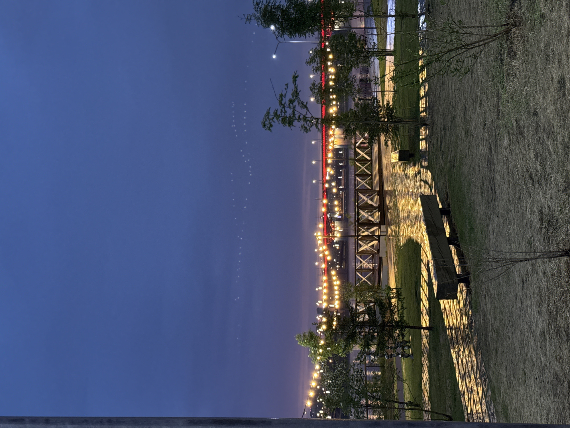 The image depicts a nighttime scene in Krasnoyarsk, Russia. In the background, a bridge is illuminated with rows of lights, including red lights along the structure. The foreground shows a park area with a bench and trees, also illuminated with lights. The sky above is a dark blue, indicating either twilight or night. The park pathway and lawn are visible, with the bench casting a shadow. The composition of the photograph is taken with a slight upward angle, showing the bridge and park in detail. A barrier appears in the bottom left.
