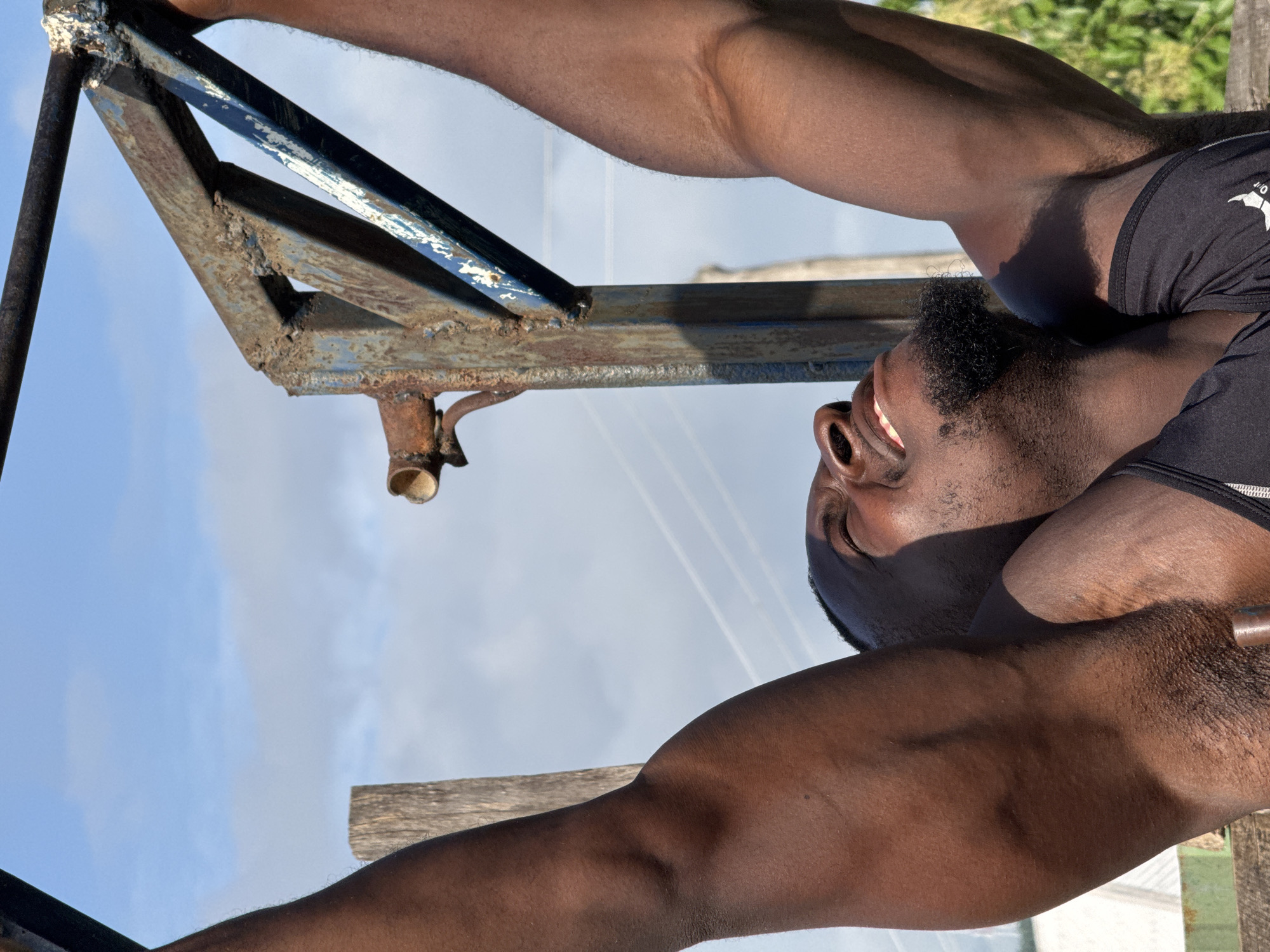 A low-angle, close-up, outdoor shot captures a Black man performing a pull-up on a rusty metal bar. The man, with a goatee and a determined expression, is sweating and looking upwards. His muscles are prominent as he grips the bar with both hands, arms extended. The background features a blue sky with scattered clouds, power lines, and a wooden post. The overall scene suggests an outdoor gym or a makeshift workout area, possibly in Akwanga, Nigeria.