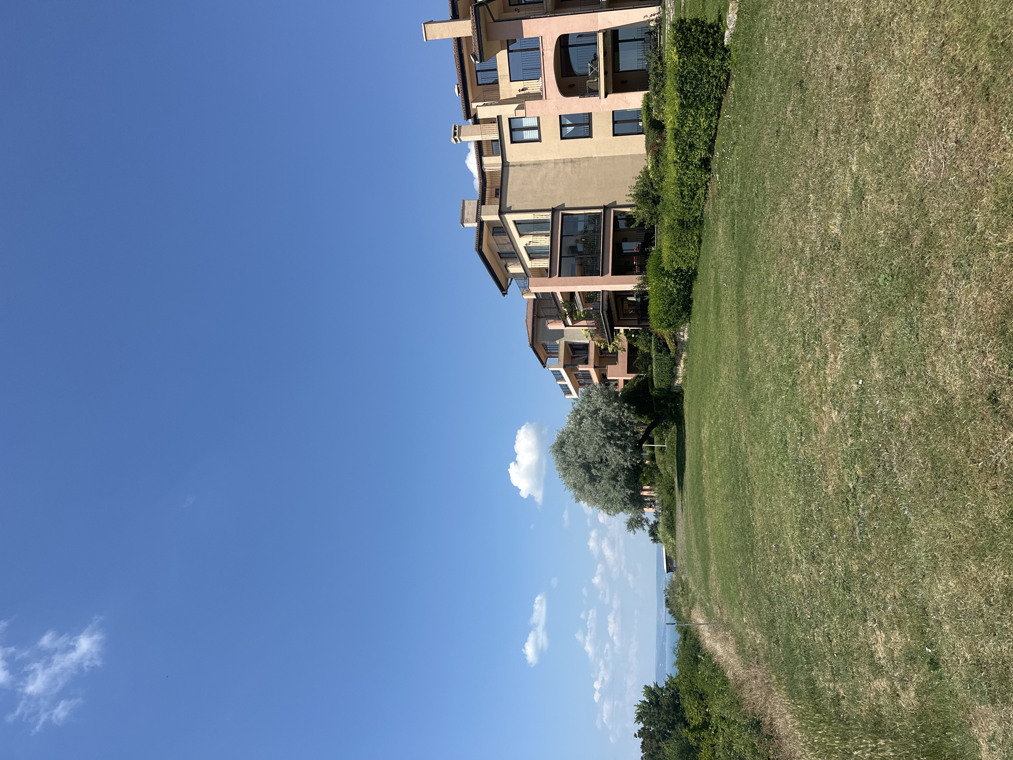 A tall, wide shot captures a row of resort-style apartment buildings along a grassy slope under a clear blue sky. The buildings, painted in shades of peach, tan, and pale pink, feature multiple balconies with dark railings and a few chimneys. They are nestled behind neatly trimmed green bushes. In the foreground, a sprawling green lawn slopes gently downward. To the left of the buildings, a large, silver-leafed tree stands prominently, its branches reaching upwards. Beyond the tree and buildings, a body of water, likely the sea, is visible under the bright sky, with a few scattered white clouds. The scene conveys a sense of a peaceful, sunny day in a coastal resort.