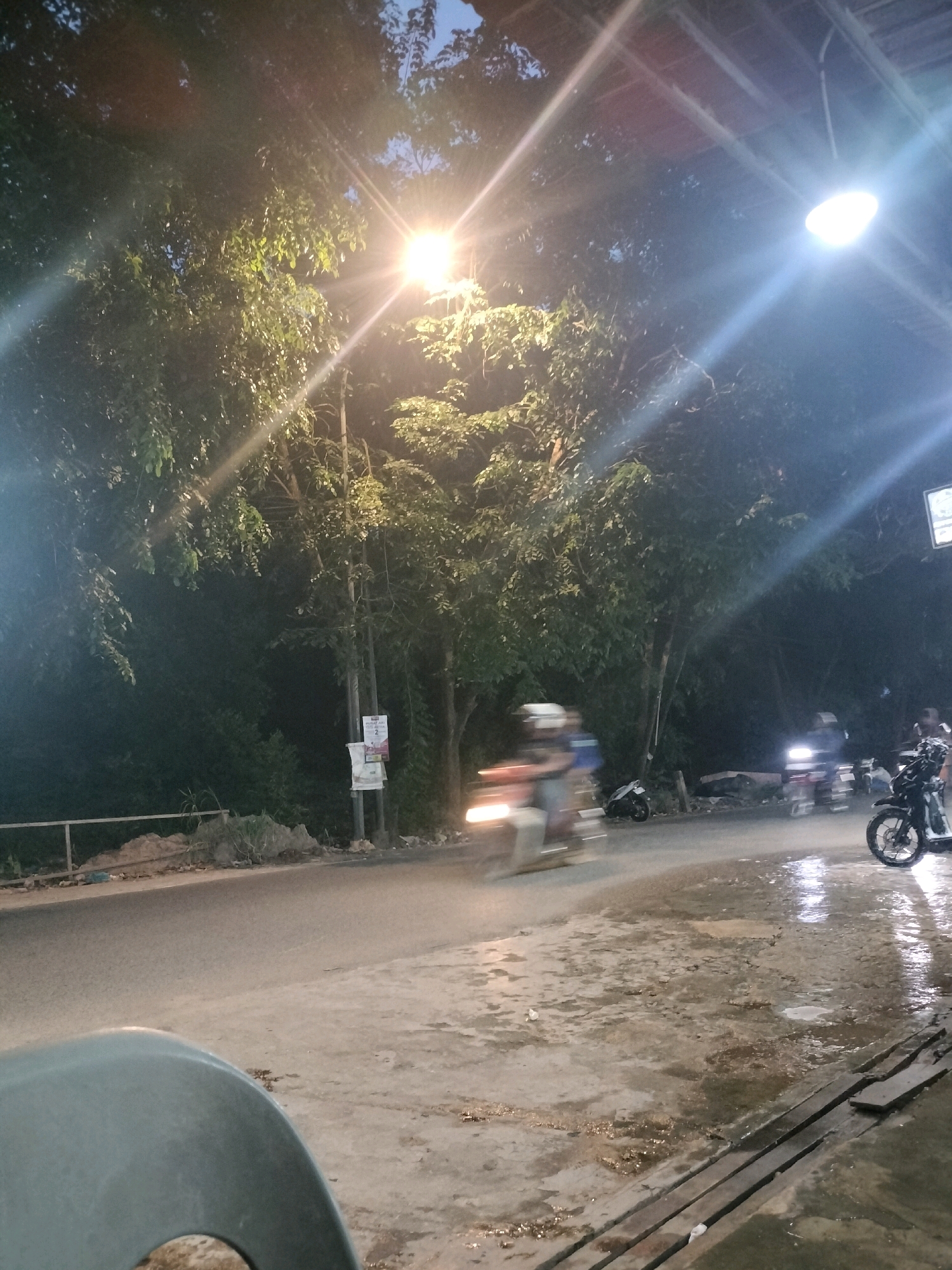 This media file captures a nighttime scene on a street in Batam, Indonesia. The primary subjects are several motorcycles and their riders, blurred due to motion. The setting appears to be a road or pathway adjacent to a line of trees. Streetlights illuminate the scene, creating lens flares and highlighting the wet ground, suggesting recent rain or ongoing dampness.
The activity depicted is the movement of motorcycles on the road. One motorcycle in the foreground is prominently blurred, indicating it's in motion. Other motorcycles and figures are visible further down the road, also appearing as streaks of light and motion. A portion of a gray plastic chair is visible in the lower left foreground, suggesting the viewpoint might be from someone sitting.
Notable details include the time of day, which is clearly nighttime due to the artificial lighting and dark sky. The wet ground and reflections of light suggest a humid or rainy environment. The presence of trees lining the road provides a natural backdrop. There is a utility pole with some posters or flyers attached, one of which has the number "2" clearly visible, along with some Indonesian text that is too small to decipher. The overall mood is one of a typical evening scene in a tropical urban or semi-urban environment.