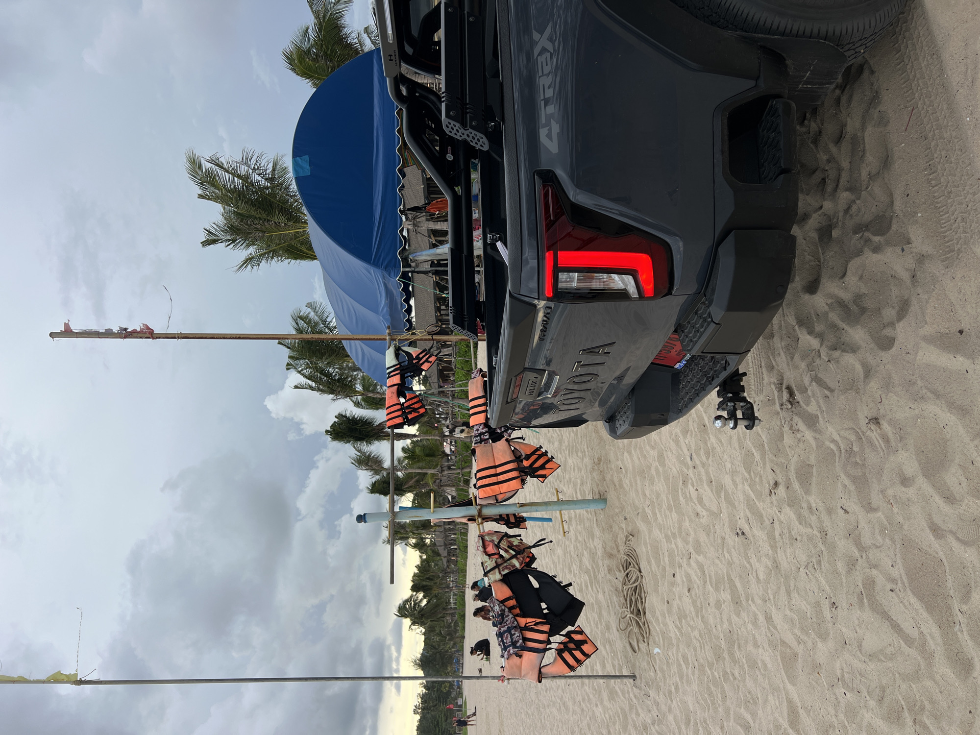 An overcast day at a sandy beach, with a grey Toyota Hilux 4x4 pickup truck parked in the foreground, facing left. The truck's rear bumper, taillights, and "4x4 TREX" badging are visible. A hitch receiver with a ball mount is attached to the truck's rear. To the left of the truck, a rack holds numerous orange and black life vests. A coiled rope lies on the sand near the rack. In the background, palm trees and a blue tent structure are present, and several people are seated on the sand. Two tall poles with tattered flags stand on the left side of the frame.
