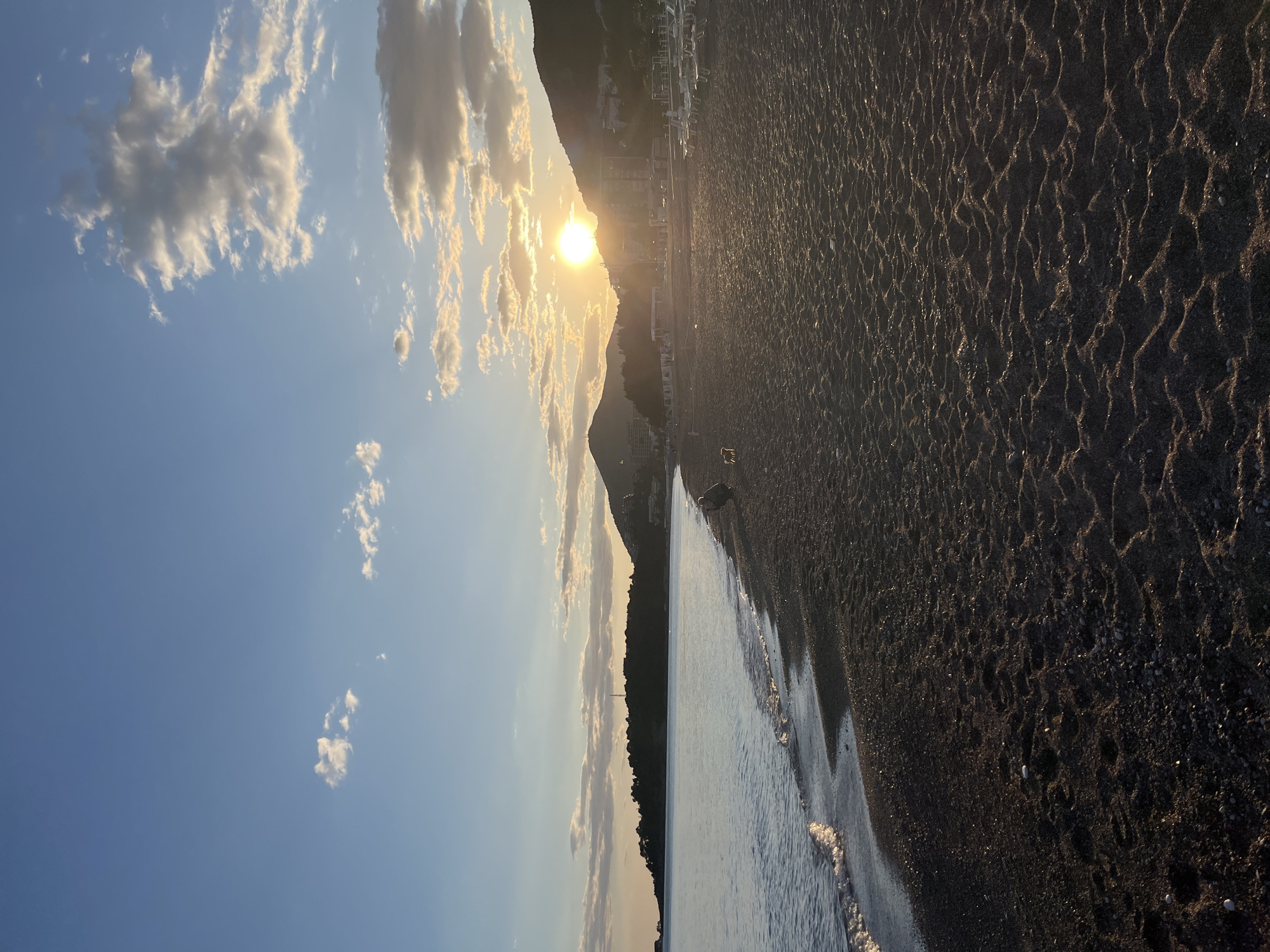 The image displays a serene coastal scene, likely at sunset or sunrise, in Rafailovići, Montenegro. Although the original file is oriented vertically, the scene is best viewed horizontally with the horizon running across the middle. The sky, occupying the upper portion of this horizontal view, is a clear blue, adorned with scattered cumulus clouds that are illuminated with warm, golden hues by the low-lying sun. The sun itself, a brilliant golden disk, is prominently featured near the center-left, partially obscured by the silhouette of a large, dark mountain.
The mountain range extends across the middle ground, its peaks and slopes cast in shadow, but with visible structures like buildings and what appear to be resort accommodations dotting its lower reaches and along the coastline in the distance. Further down the coast, some white sunbeds or umbrellas are faintly discernible on a brighter patch of beach. The right side of the image is dominated by a dark, textured beach, likely composed of pebbles or coarse sand. The sand exhibits distinct ripple patterns, possibly formed by water or wind, with small, lighter-colored stones scattered intermittently across its surface.
Along the very bottom of the frame, the gentle waves of the sea meet the shore, creating a thin line of white foam. The water appears calm, reflecting the soft light of the sky. In the mid-ground, near the water's edge on the beach, a very small and indistinct dark shape is visible, possibly a distant person or animal, but it is not clearly identifiable. The warm, golden light casts long shadows, enhancing the tranquil atmosphere of the beach at either dawn or dusk. No visible text is present in the image.