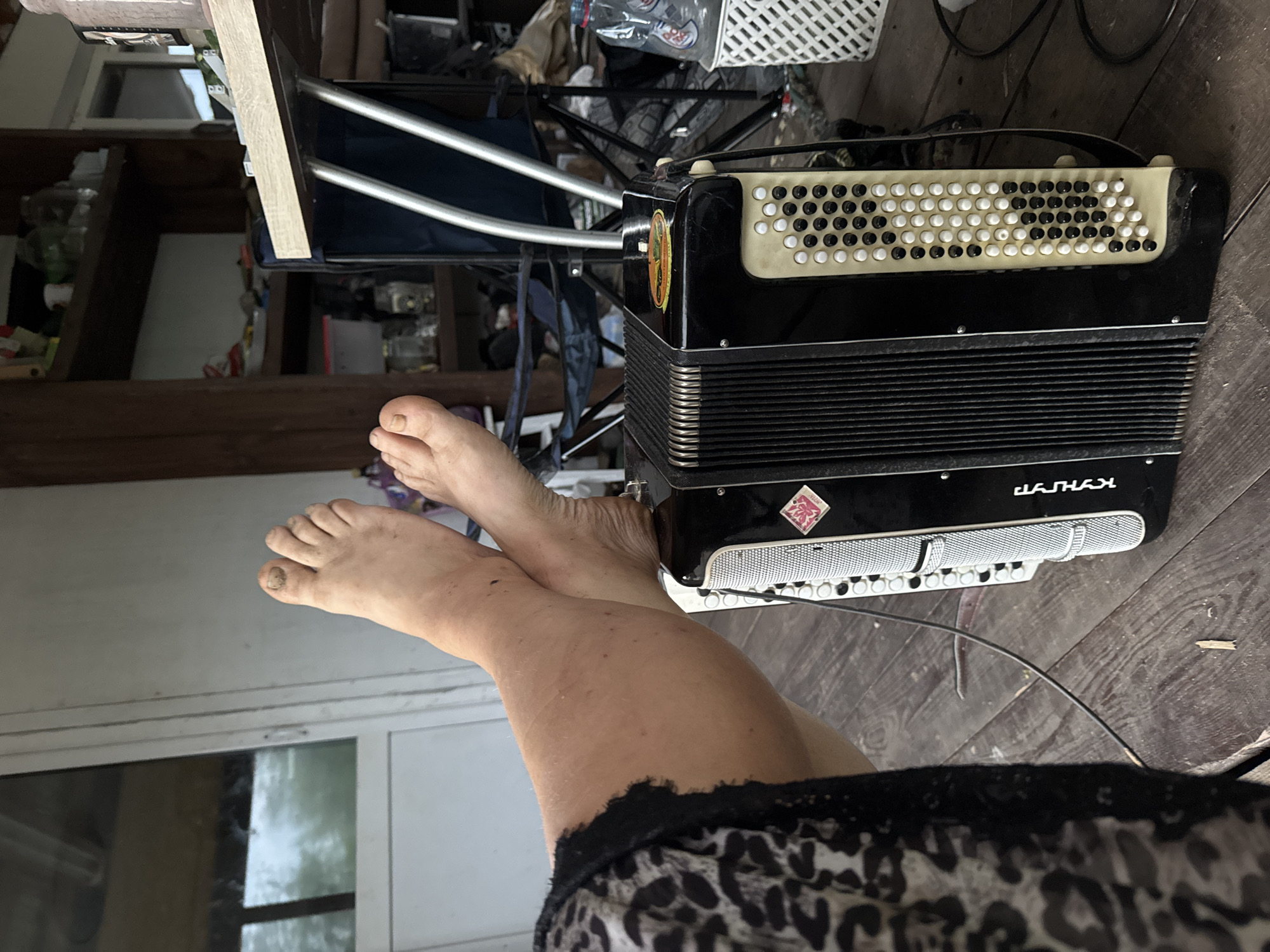 In the city of Chaikovsky, Russia, a person's bare feet are resting on a black accordion. The feet are dirty and appear to be tan. The person is wearing a black and white leopard print skirt or dress that covers their legs. The accordion is on a wooden floor, possibly a porch or deck. In the background, there are shelves filled with various items, a blue folding chair, and a white plastic basket with bottles. The overall environment suggests an outdoor or semi-outdoor setting.