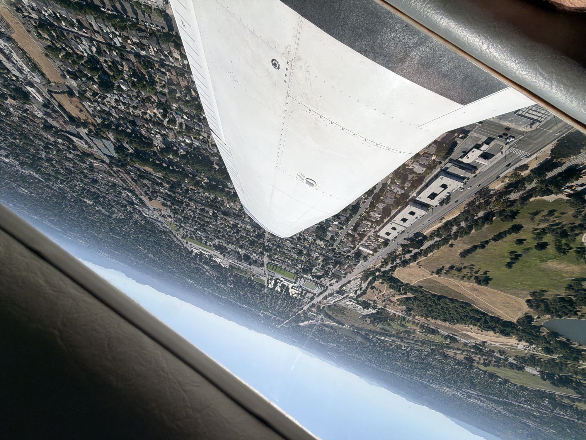 The image shows an aerial view from a small airplane, likely during a turn. The wing of the aircraft is prominent in the foreground. Below, the landscape features a dense urban area with residential and commercial buildings, roads, a large park or golf course, and a distant mountain range obscured by haze. A football stadium and a secondary school can be identified. It is over Berkeley, CA.