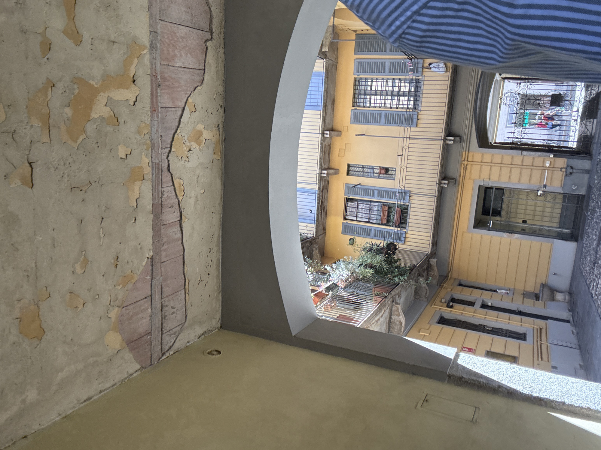 The image captures a view of a courtyard in Milan, Italy, looking through an archway. The courtyard features yellow buildings with balconies and shuttered windows. In the foreground, the underside of an archway is visible, with peeling paint and exposed brickwork on the ceiling. To the right, a person wearing a blue and white striped shirt is partially in frame. Through the archway, a decorative wrought-iron gate leads to another area with people visible. The architecture suggests an older urban setting.