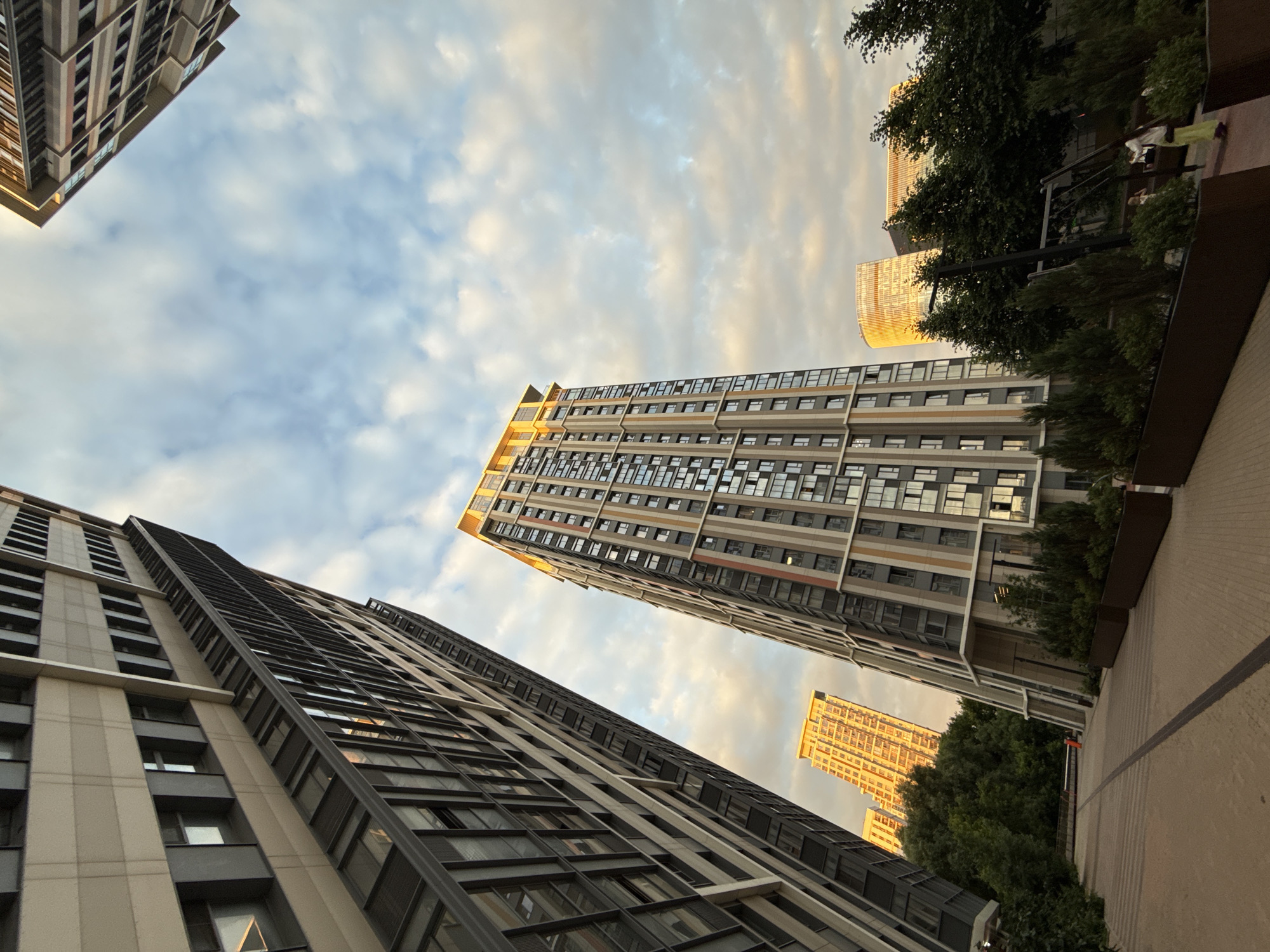 The image depicts a modern urban scene with towering residential buildings set against a cloudy sky during what appears to be either sunrise or sunset, indicated by the warm golden light reflecting off the glass facades of some structures and the soft hues in the clouds. The perspective is from a low angle, looking upwards, which emphasizes the scale and height of the architecture.
The primary subjects are the buildings themselves, characterized by their repetitive window patterns and contemporary design. Several buildings are visible, with varying architectural styles and colors, suggesting a relatively new development. One building in the mid-ground stands out with its warm, golden illumination, possibly due to the angle of the sun. Another taller building on the left is rendered in shades of beige and dark gray, with a multitude of windows. In the background, more buildings and lush green trees can be seen, adding a touch of nature to the urban landscape.
The scene appears to be a residential complex or a mixed-use development. There are no people prominently featured in the image, though glimpses of figures might be present within the architecture or in the background, but they are not the focus. The activity, if any, is not discernible, suggesting a moment of quiet observation rather than an active event.
Notable details include the soft, diffused light that creates a serene atmosphere, and the interplay of shadows and light on the buildings. The cloudy sky suggests a dynamic weather condition. The presence of trees indicates some landscaping or park areas within or adjacent to the buildings. The location context of Moscow, Russia, is given, which can be inferred to align with the typical modern high-rise architecture found in large Russian cities. No visible text is present in the image.