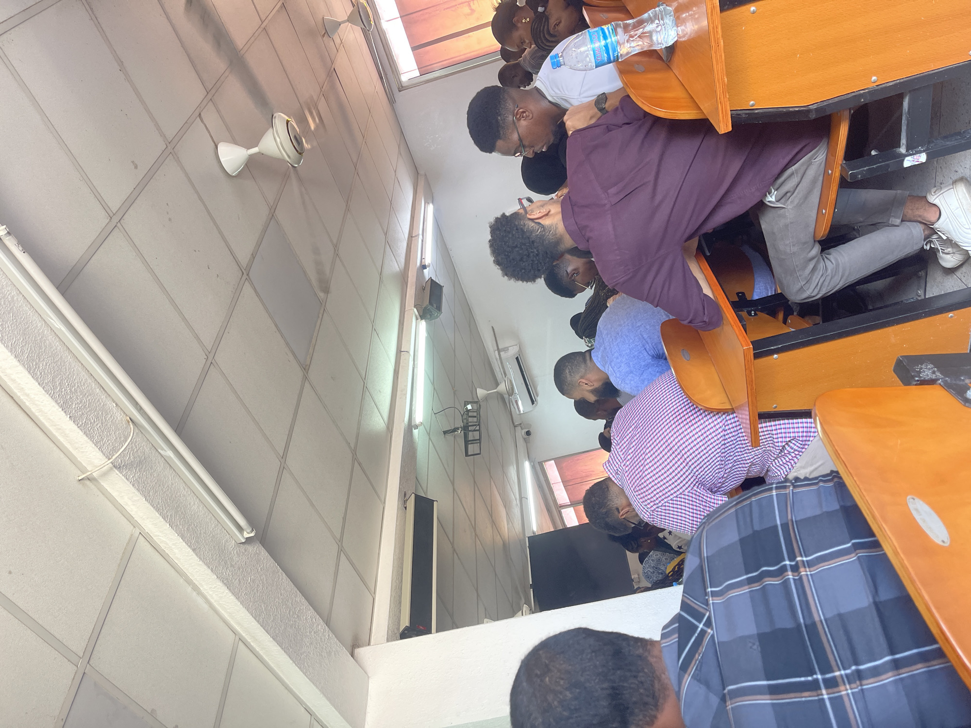 The image depicts an indoor scene, likely a classroom or lecture hall, with rows of students seated at desks. The students are of various ethnicities and appear to be in their late teens or early twenties. They are dressed in casual attire. The desks are wooden and have attached seats. A water bottle is visible on one of the desks. The walls of the room are painted white and have fluorescent lighting fixtures and ceiling fans. There are windows visible in the background. The location is not explicitly identified, but the prompt suggests it might be Dar es Salaam, Tanzania.