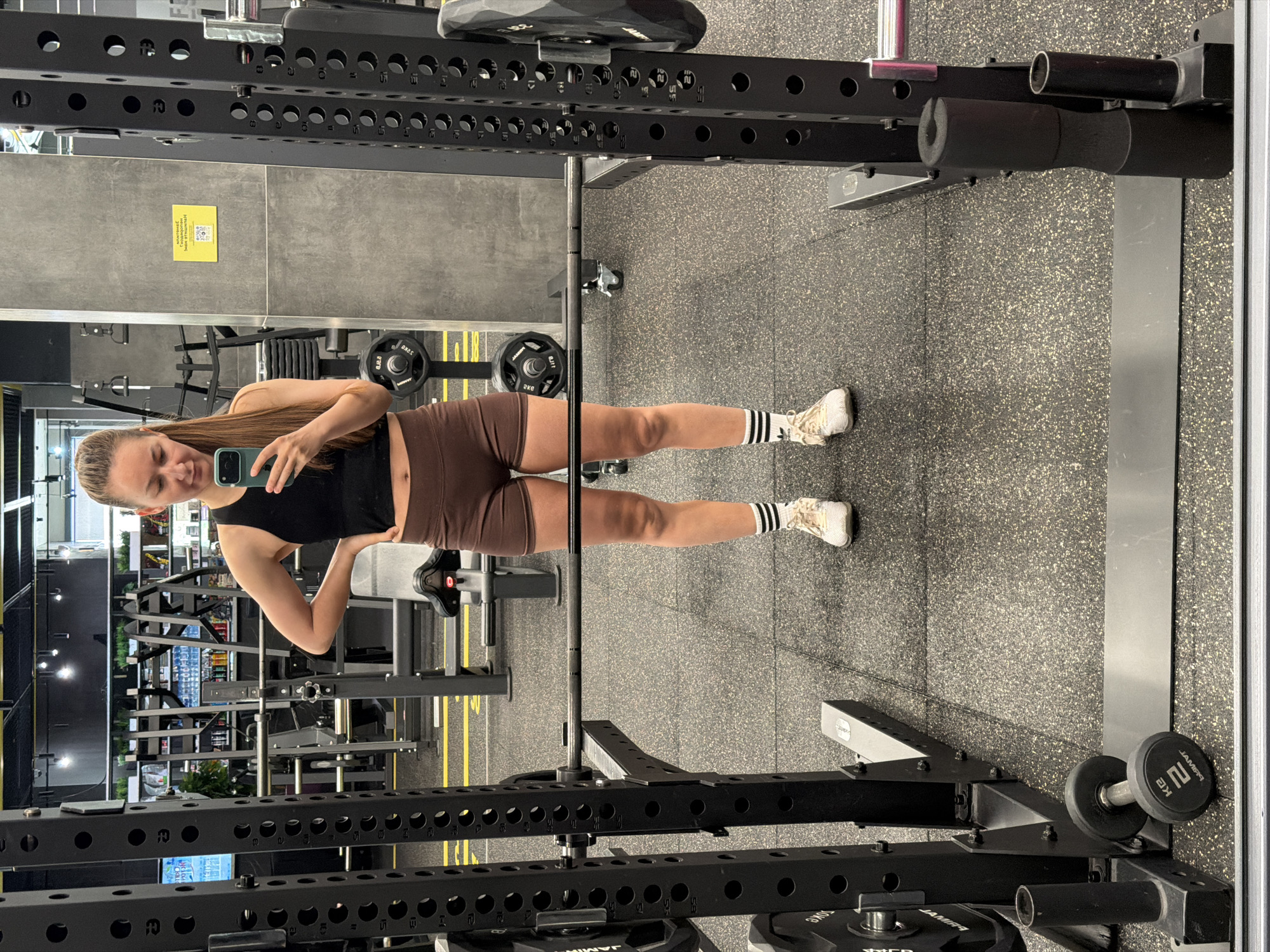 A young woman is seen taking a selfie in a gym setting. The gym appears to be located within the city of Tyumen, Russia as per the information provided.
The woman is wearing a black tank top and brown shorts. She is standing between a weight rack and a mirror. In the background, gym equipment such as weight machines and racks are visible. The floor is covered in a black rubber mat. A mirror is present along the right side of the image.