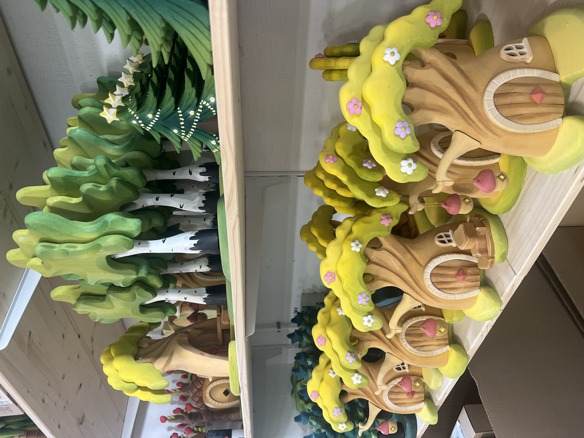 The image shows a shelf filled with handmade wooden toys. On the top shelf, there are several stylized wooden trees with green leaves and white trunks, and a decorated Christmas tree with yellow stars. On the lower shelf, there are multiple whimsical treehouse-like structures, each with a rounded door, a small window, and decorative pink and white flowers on the yellow canopy. Some of these treehouses have pink acorn-shaped lanterns hanging from them. The toys are arranged neatly on wooden shelves. The environment appears to be a workshop or a retail display.