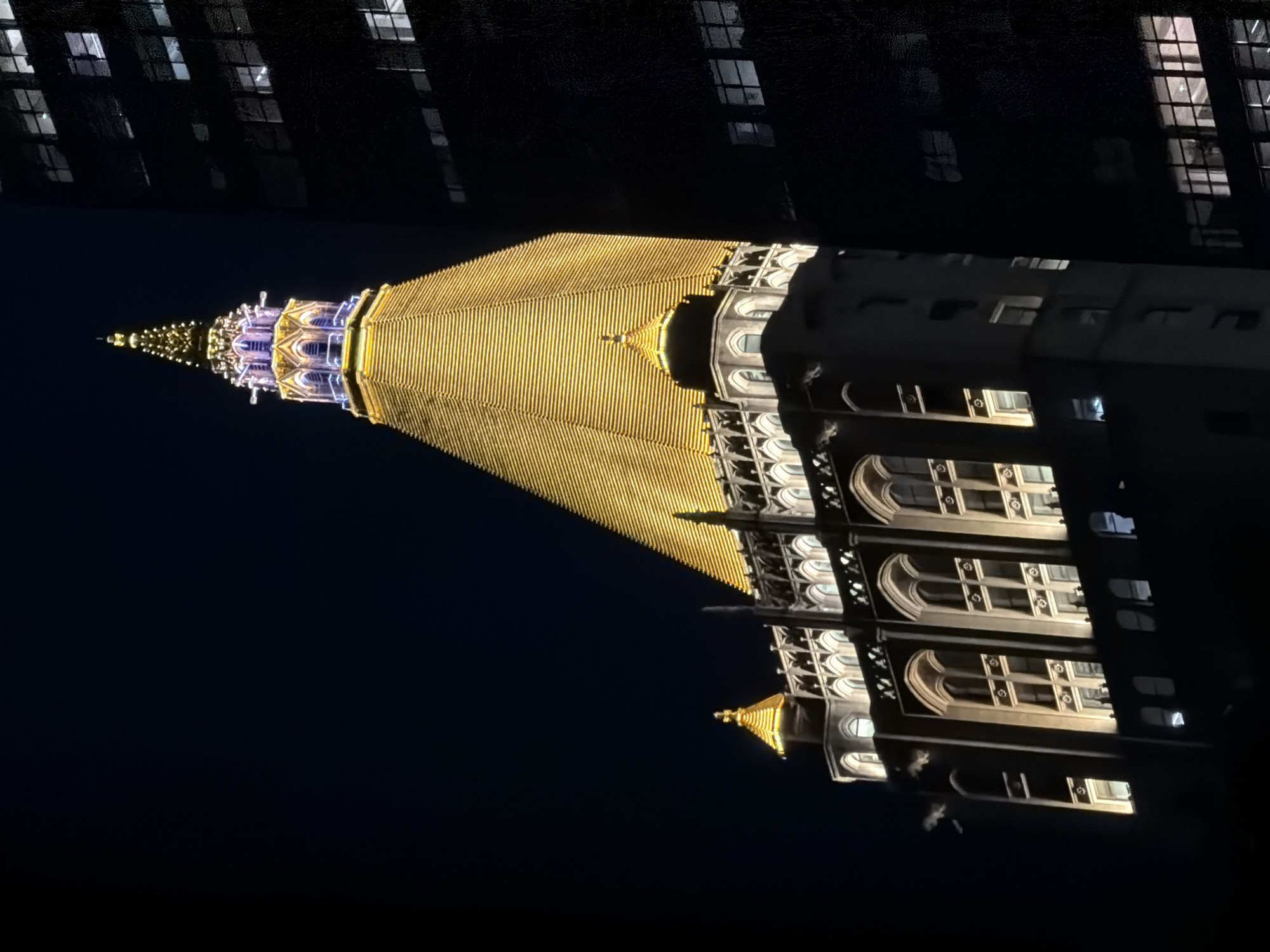 This is a night time, low angle shot of the Starrett-Syracuse building, a historic skyscraper in Midtown Manhattan, New York City, United States of America, illuminated by warm, golden lights. The building's distinctive tiered, pyramid-like roof is prominently featured, topped by an ornate spire with blue and gold accents. Gothic-style architectural details are visible on the lower levels of the building, with arched windows and decorative stonework. To the right of the Starrett-Syracuse building, a darker, modern skyscraper looms, its windows reflecting the ambient light. The sky is a deep, dark blue, indicating it is nighttime. There are no people or discernible animals visible in the frame.