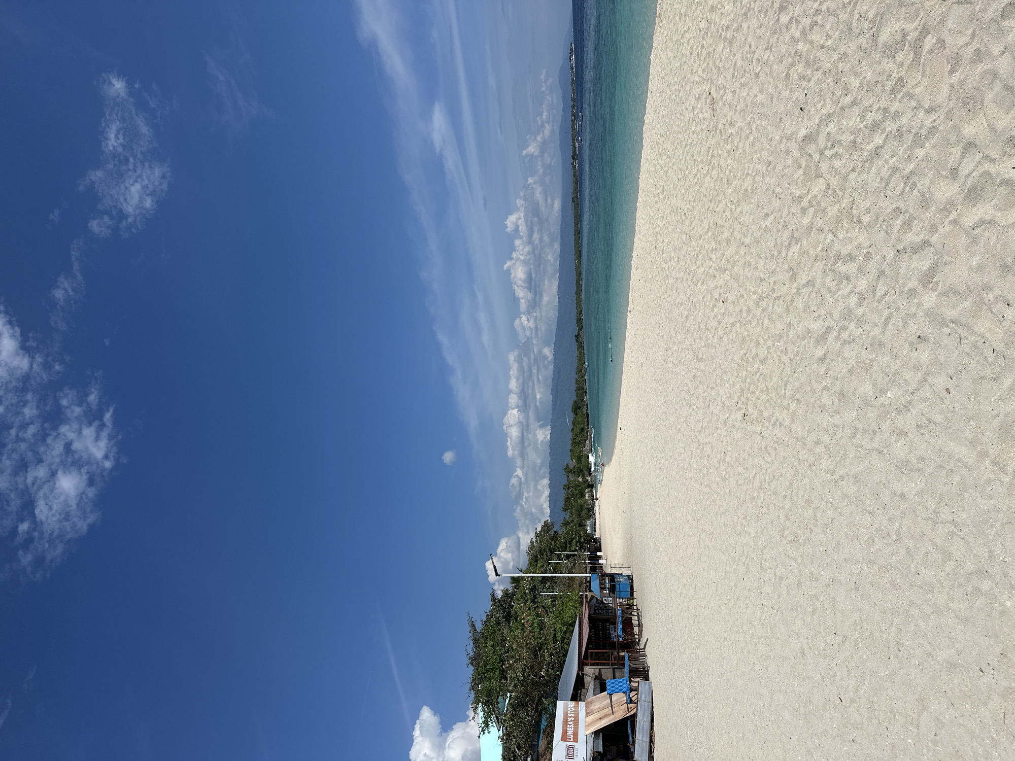 The image depicts a pristine white sand beach on a sunny day, likely in Saavedra, Philippines, based on the provided context. The foreground is dominated by the expansive sandy beach, showing subtle undulations and scattered small debris. In the middle ground, to the left, a small cluster of structures appears to be commercial establishments or stalls. One structure, identified by signage as "TANGUY LUNESA'S STORE," has a rustic appearance with wooden planks and a covered area. Blue chairs are visible outside this store. Beyond these structures, a line of lush green trees runs along the edge of the beach. A street lamp stands near the trees.
The turquoise water of the sea stretches out to the horizon, with a clear demarcation between the shallow, lighter blue near the shore and the deeper blue further out. Several small boats are scattered on the water. In the distance, a range of mountains forms the coastline, partially obscured by clouds. The sky is a vibrant blue, dotted with fluffy white clouds, some of which are thin and wispy, while others are more substantial cumulus formations. The overall atmosphere is tranquil and idyllic, characteristic of a tropical beach destination. A few indistinct figures are visible walking on the beach and swimming in the water.