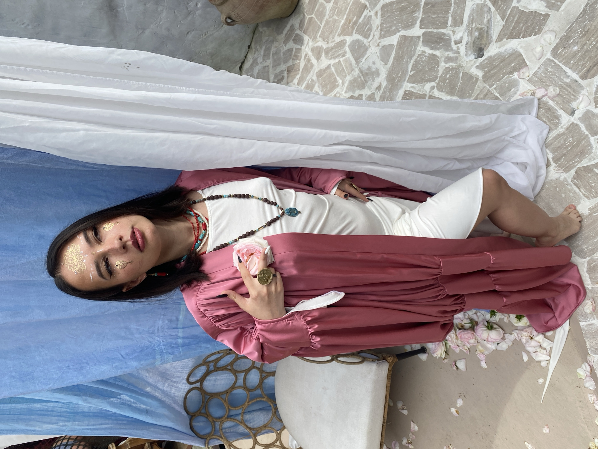 A young woman with dark hair and gold face paint stands indoors, partially obscured by white and blue fabric draping. She is wearing a flowing pink robe over a white dress and multiple beaded necklaces. In her right hand, she holds a pink rose and a ring with a geometric design. The floor is paved with irregular stones, and rose petals are scattered around. A white upholstered chair with a decorative metal frame is visible to her left, and a large ceramic pot is to her right. The scene appears to be a studio or outdoor setting. The woman's posture suggests a contemplative or posed moment. The overall aesthetic is bohemian or mystical. The location is not specified as Sochi, Russia.