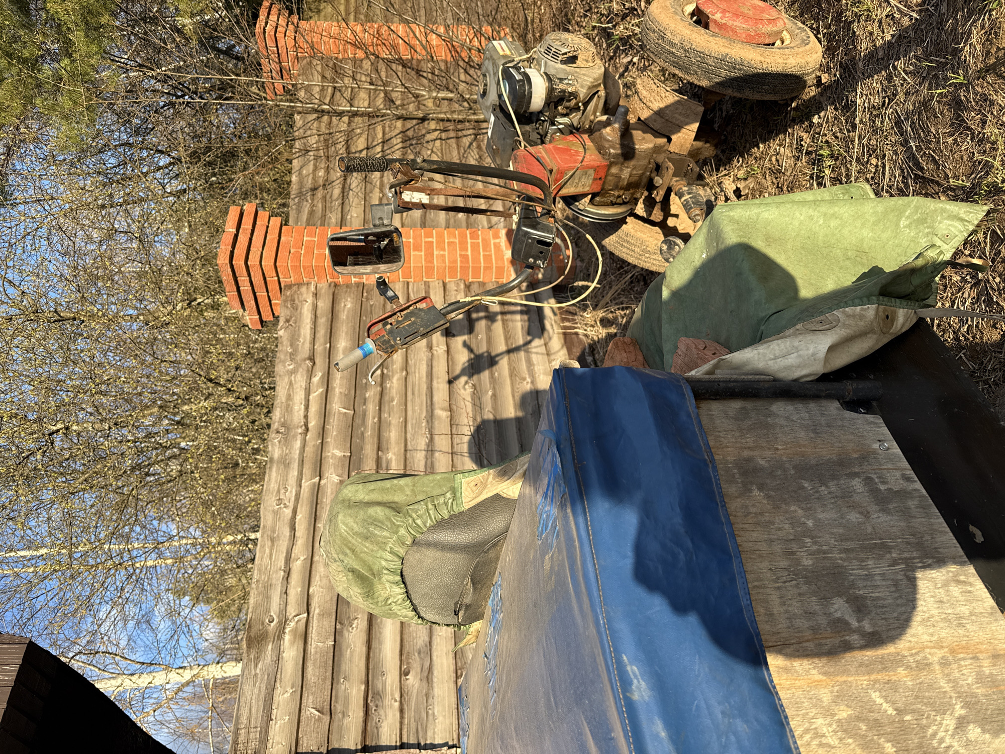 The image shows an outdoor scene, possibly a backyard or storage area, in Galevo, Russia. The environment appears rustic, with a wooden fence and ground covered in dirt and some vegetation.
Several objects are present: a gasoline-powered engine, possibly from a tiller or other small machine, rests on a small wheeled chassis, bricks, and what looks like a blue tarp or protective cover. There is also a smaller motorized machine.
No people or interactions are visible in the image.