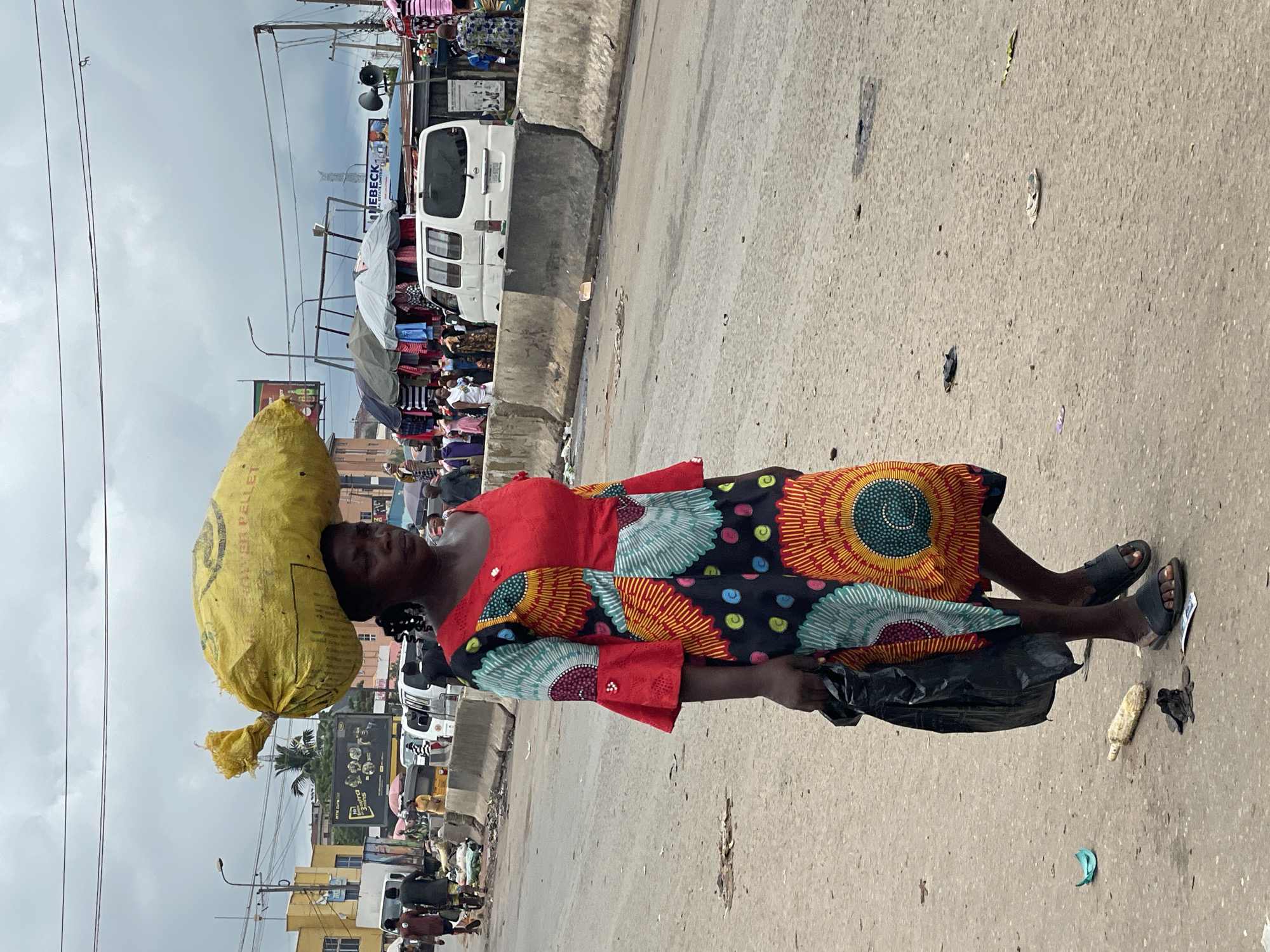 A Black woman with a large yellow bag on her head walks down a paved street in what appears to be a market or busy commercial area. She is wearing a brightly colored, patterned dress with bell sleeves and black sandals. In her left hand, she carries a black plastic bag. The street is lined with concrete barriers, market stalls with hanging clothes, and various vehicles, including a white van. The ground is littered with debris, including trash and what looks like discarded food. The sky is overcast with gray clouds. The background shows buildings and power lines.