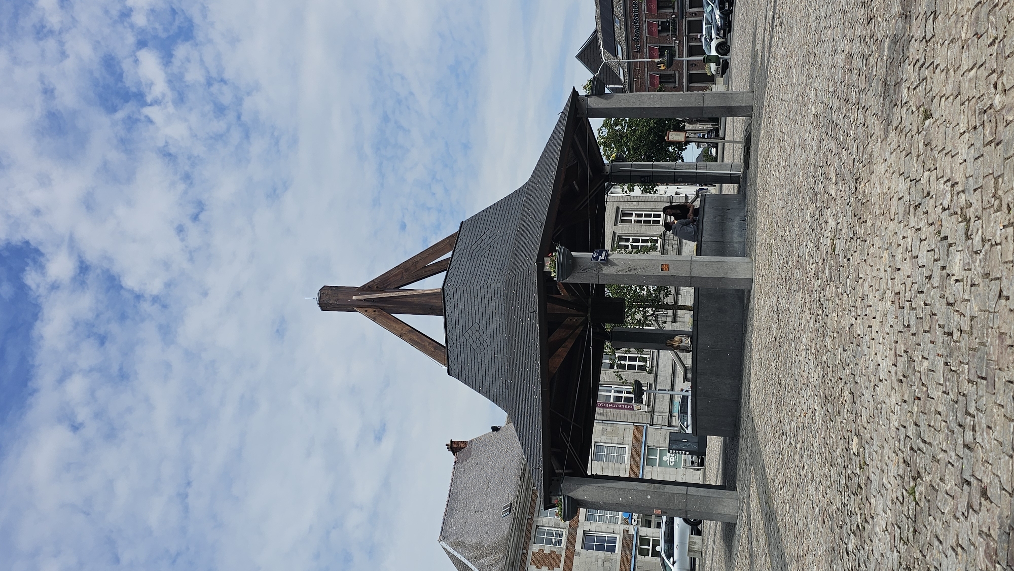 A well structure is visible in the center of the image, in the Place des Trois Fers in Philippeville, Belgium. The well structure has a dark grey, hexagonal roof with a wooden apex structure. The roof is supported by four thick, grey stone pillars. Inside the well structure, a grey stone basin is visible. There are two people sitting on the edge of the basin, one with blonde hair and the other with dark hair. They are facing away from the camera, towards the well. Behind the well structure, there are buildings with traditional architecture. The ground in front of the well is paved with cobblestones. The sky above is blue with scattered white clouds. The overall impression is of a peaceful town square on a cloudy day.