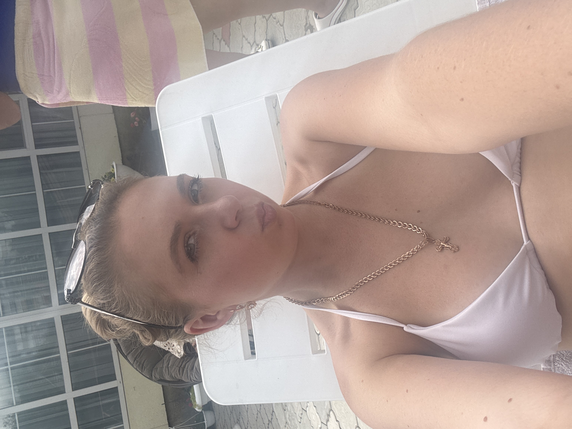 A selfie is taken outdoors by a young woman wearing a light-colored bikini. She is lying on a white lounge chair, and her upper body is visible. Her blonde hair is pulled back, and she is wearing sunglasses perched on her head. A delicate gold chain with a cross pendant rests on her chest. The background shows a glimpse of a building with white-framed windows and a patio area. The lighting suggests it is daytime, possibly sunny. The woman's expression is a pout, and she appears relaxed and possibly posing for the camera. The image seems to capture a casual moment, likely during a vacation or a warm weather day.