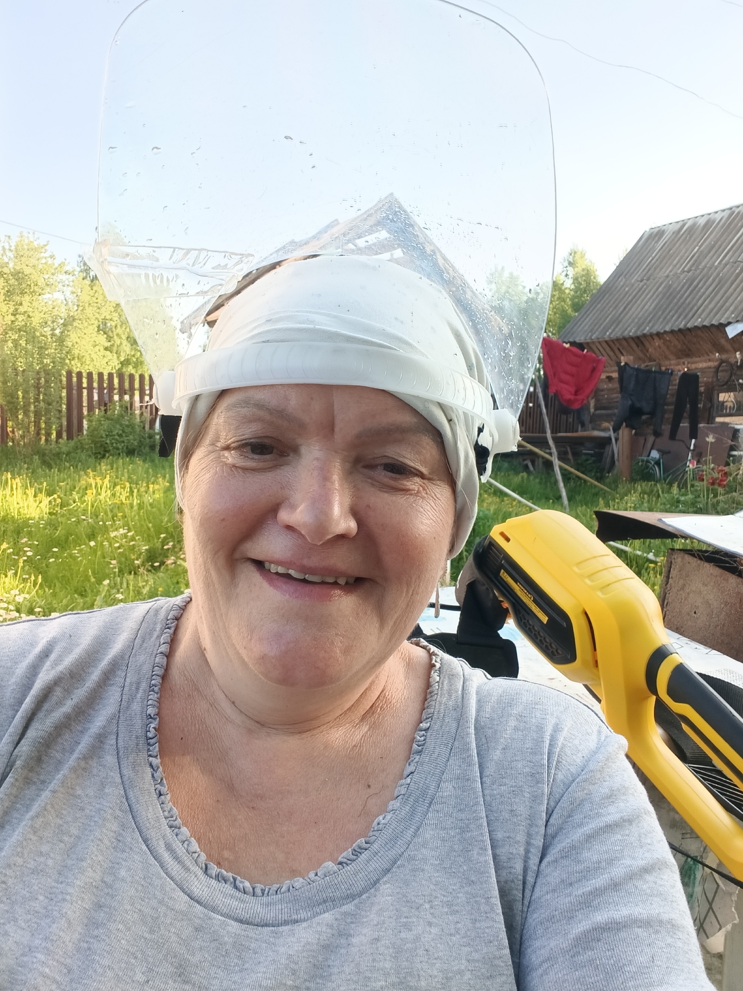 A smiling middle-aged woman in Красный Маяк, Russia, wears a light gray t-shirt with a ruffled neckline and a white head covering with a transparent face shield. She is holding a yellow and black handheld device, possibly a tool or equipment. The background includes a rural landscape with a wooden fence, greenery, a wooden building with clothes hanging on a line, and miscellaneous objects. The lighting suggests it is daytime.