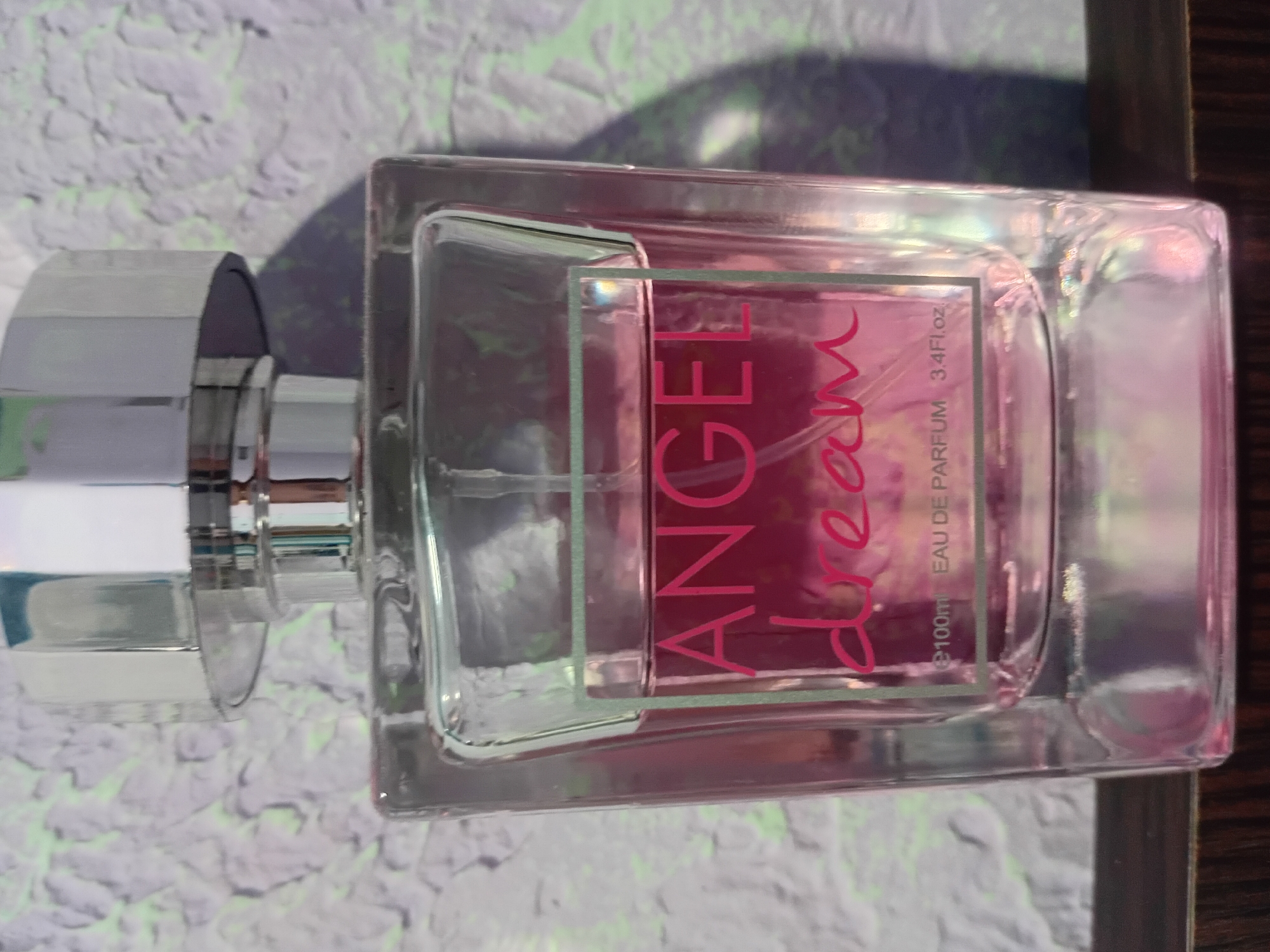 A square glass perfume bottle with a silver cap sits on a dark wooden surface. The bottle is filled with a pink liquid and has a label that reads "ANGEL dream" in pink font. Below this, it says "e100ml EAU DE PARFUM 3.4Fl.oz". The background is a textured purple and white wall. There is no discernible indication of the location in Bocani, Moldova.