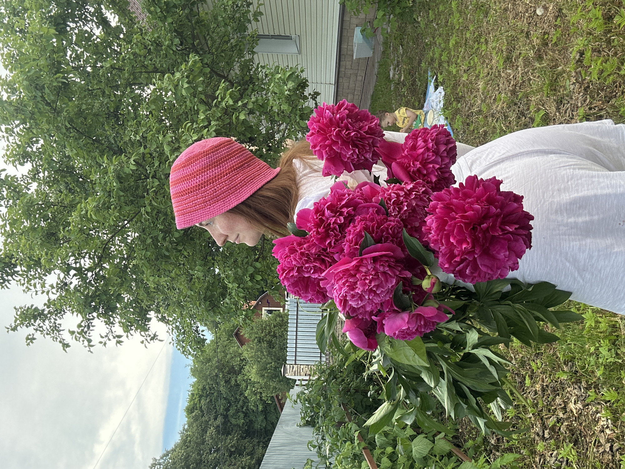 A young woman with red hair and freckles, wearing a pink striped beanie and a white shirt, stands in a grassy, suburban yard. She is holding a large bouquet of vibrant pink peonies. Behind her, a fence encloses the yard, and in the distance, a house with light siding and a dark roof is visible. A small child in a yellow shirt sits on a blanket in the background. The environment appears to be a backyard in Nizhny Novgorod, Russia, with lush green trees and a cloudy sky.