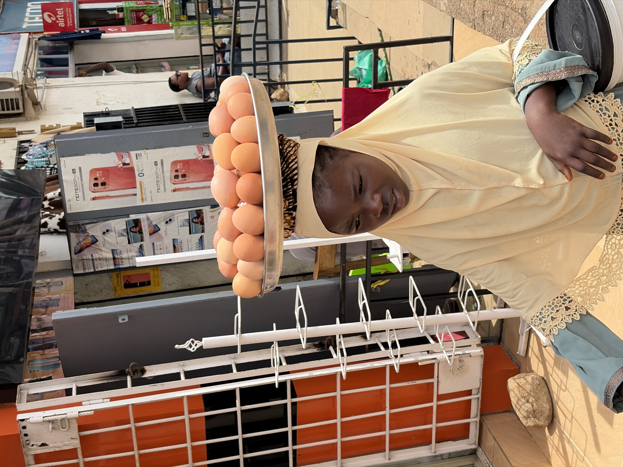 The image depicts a young girl in Jos, Nigeria, wearing a light yellow hijab and a blue-green dress with intricate lace trim. She is carrying a large metal tray filled with numerous eggs balanced on her head, supported by a woven mat. The girl's gaze is directed downwards and to her left, with her right hand resting near her chest. In the foreground, a black plastic bucket with a white rim is visible.
The background reveals a street scene with various elements. To the left, a white metal gate with orange bars is prominent, adjacent to a building entrance with a gray door. Further back, posters for mobile phones, including "NOTE60 PRO 5G", are displayed on a wall. A man wearing sunglasses and a grey sleeveless shirt is seated in the middle ground. The street itself is paved with tiles, and a large stone rests on the ground to the left. An Airtel advertisement can be seen on a building in the distance. The overall environment suggests an outdoor market or commercial area in a Nigerian city.