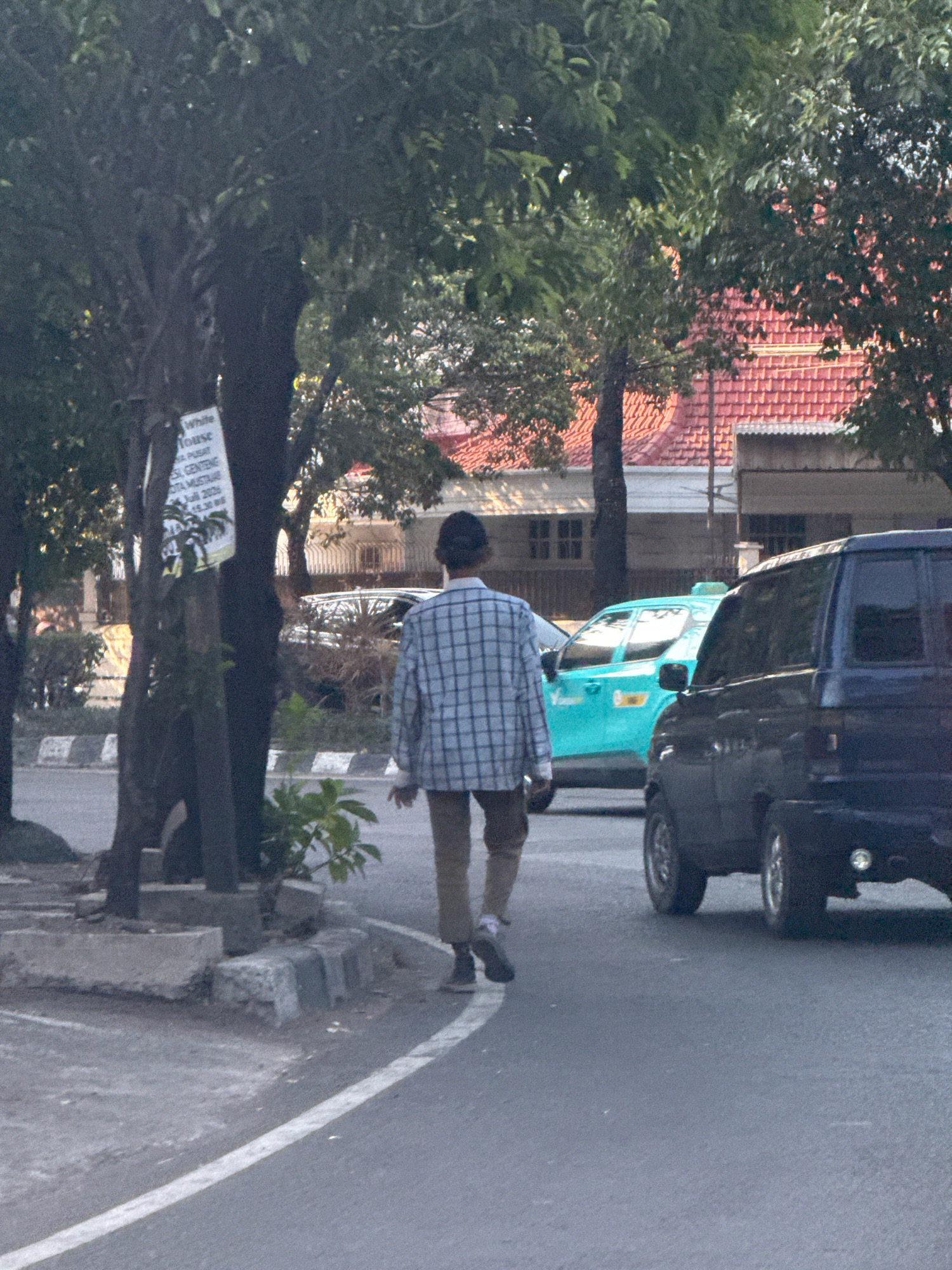 The image shows a street scene in Surabaya, Indonesia. A person wearing a checkered shirt and tan pants is walking away from the camera on the right side of the road. To the left of the person, a teal-colored taxi is parked. Further to the left, a dark-colored SUV is also parked. The background features buildings with red tiled roofs and trees. A sign attached to a tree on the left has some text, but it is mostly illegible. The road has a white line marking and a sidewalk made of concrete blocks. The lighting suggests it is daytime.