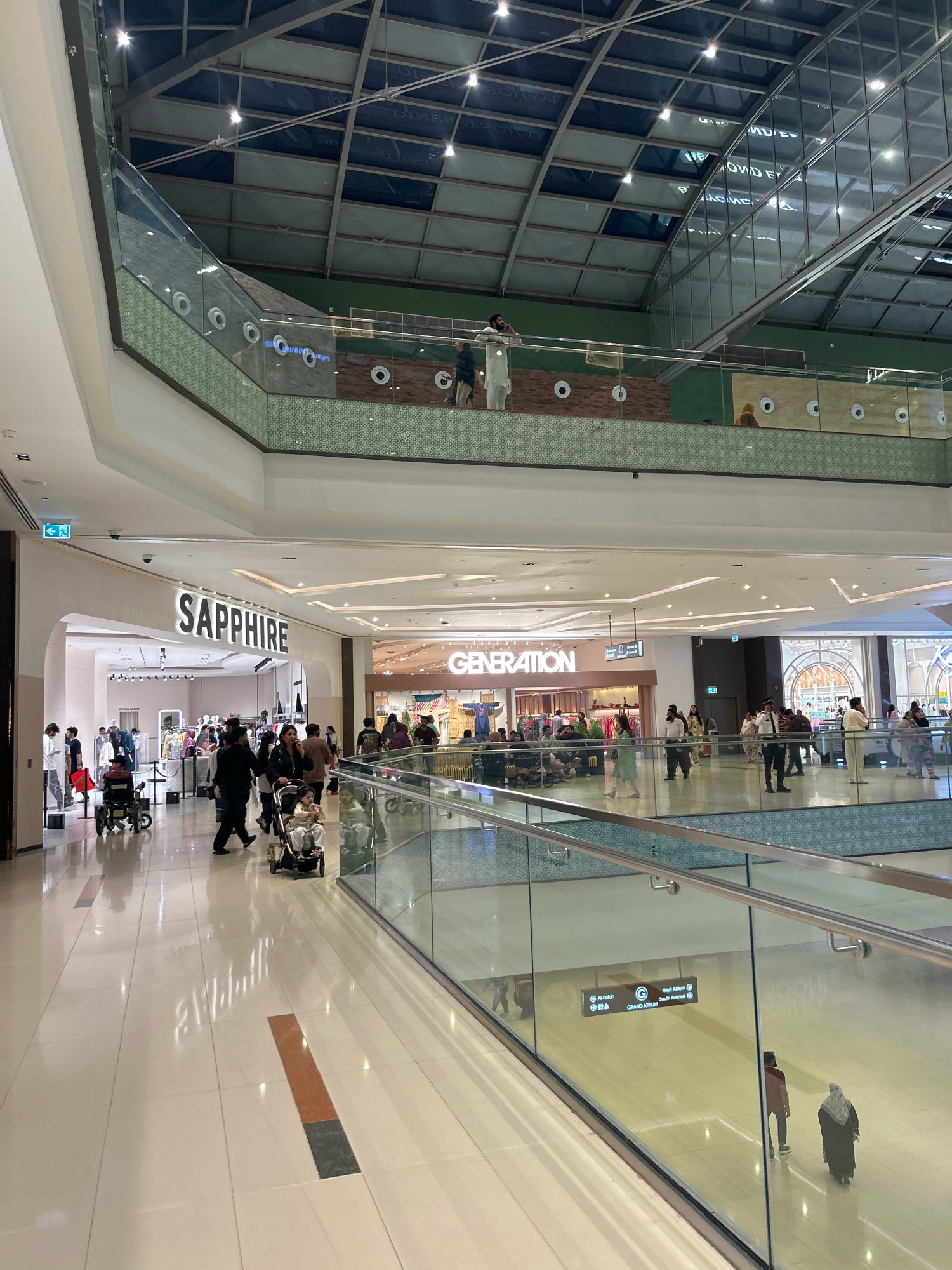 The image depicts the interior of a modern, multi-level shopping mall. The architecture features a high, vaulted ceiling with a glass and metal roof structure, allowing natural light to filter in. Various shops are visible at different levels, including "SAPPHIRE" and "GENERATION," indicated by prominent signage. The mall's interior is spacious with polished floors and glass balustrades on the upper levels overlooking the main floor.
On the ground floor, several people are walking, some pushing strollers. There is a person in a wheelchair and another individual using a shopping cart. A security guard is standing near the "GENERATION" store. People are dressed in casual and traditional attire, including some wearing hijabs. The overall atmosphere suggests a busy retail environment.
The scene shows a well-lit, clean, and contemporary shopping complex. The location is identified as the city of Chachowali, Pakistan.