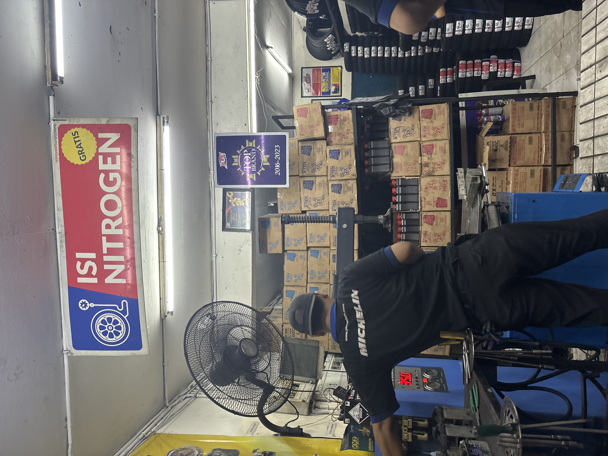 The image depicts the interior of a tire shop in North Jakarta, Indonesia. In the foreground, a man wearing a black shirt with "MICHELIN" printed on the back is operating a blue tire inflation machine. He is facing away from the camera. Behind him, shelves are stocked with boxes of various products, including what appear to be tire-related items and fluids. The shop features fluorescent lighting, a large black oscillating fan mounted on the wall, and a prominent sign above the entrance that reads "ISI NITROGEN GRATIS" (Nitrogen Fill Free) with an image of a tire and a pressure gauge. Another sign displaying "TOP BRAND 2016-2023" is visible on a shelf. Stacks of tires are visible in the background. The overall environment suggests a service-oriented business.