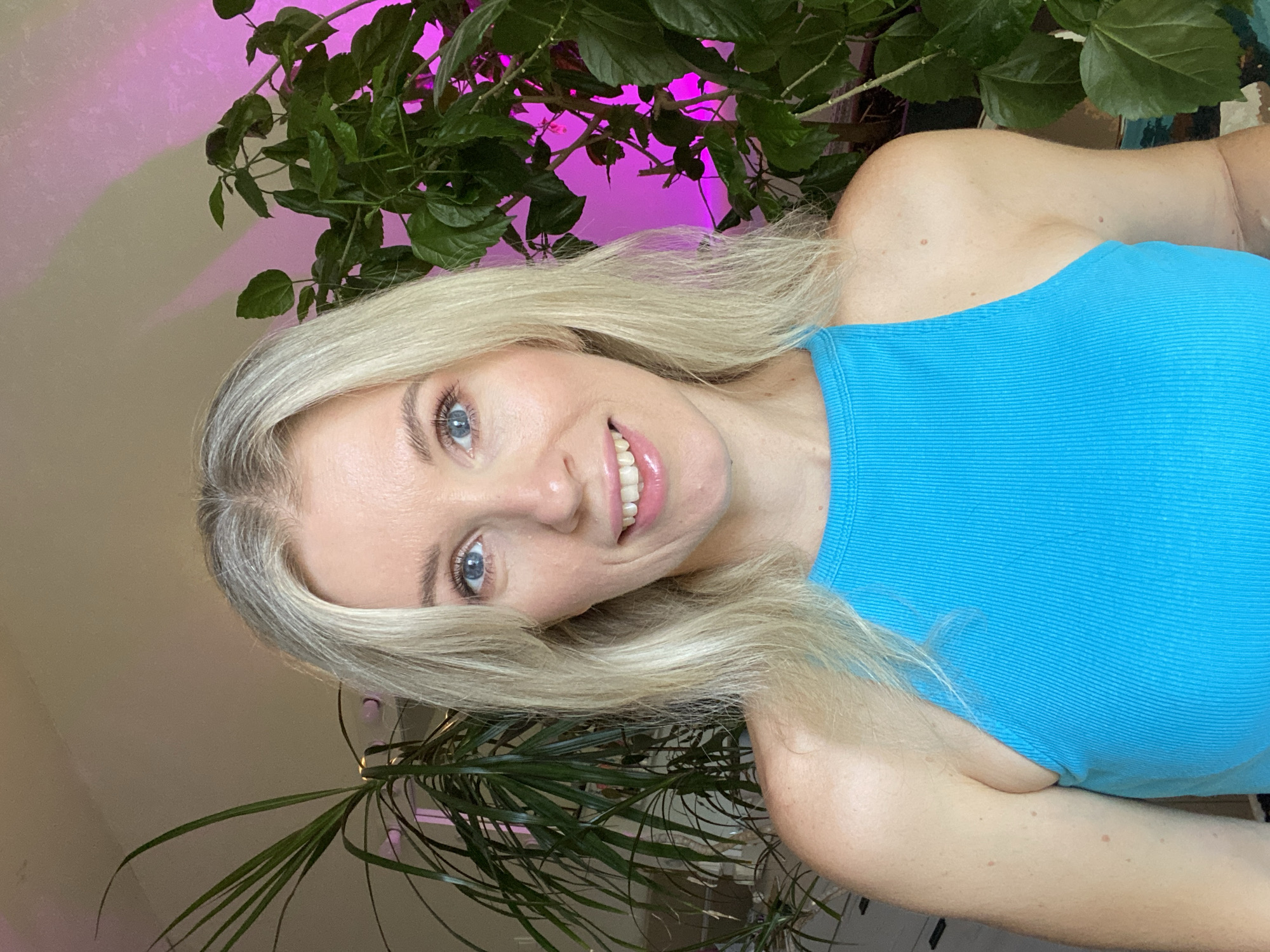 A young Caucasian woman with blonde hair and blue eyes is pictured from the chest up, smiling and looking at the camera. She is wearing a bright blue sleeveless top. Behind her, to her left, is a large leafy green plant with slender leaves. To her right is another large leafy green plant with wider leaves, illuminated by a pink light. The background is a plain, light-colored wall. The scene appears to be indoors, likely in a home or studio.