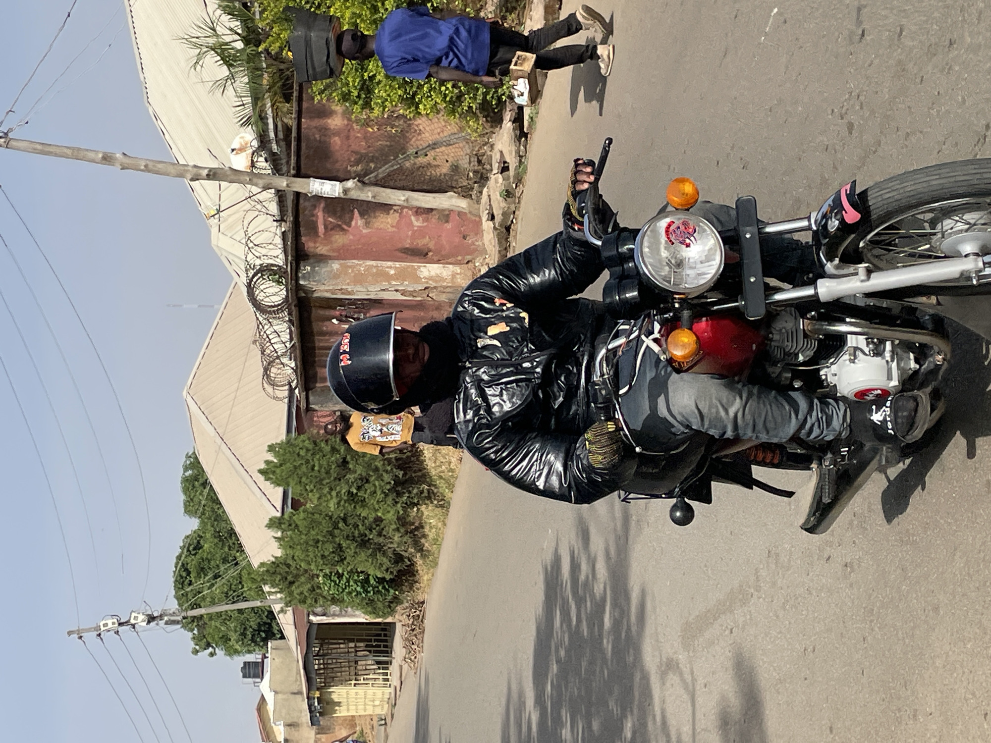 A man in a black jacket and helmet is riding a motorcycle on a city street in Unknown, Unknown. The street appears to be paved and runs along a row of houses and other structures. Another person wearing a blue shirt and black pants stands on the side of the road. Powerlines extend along the left side of the road, and there is a patch of trees on the side of the road as well.