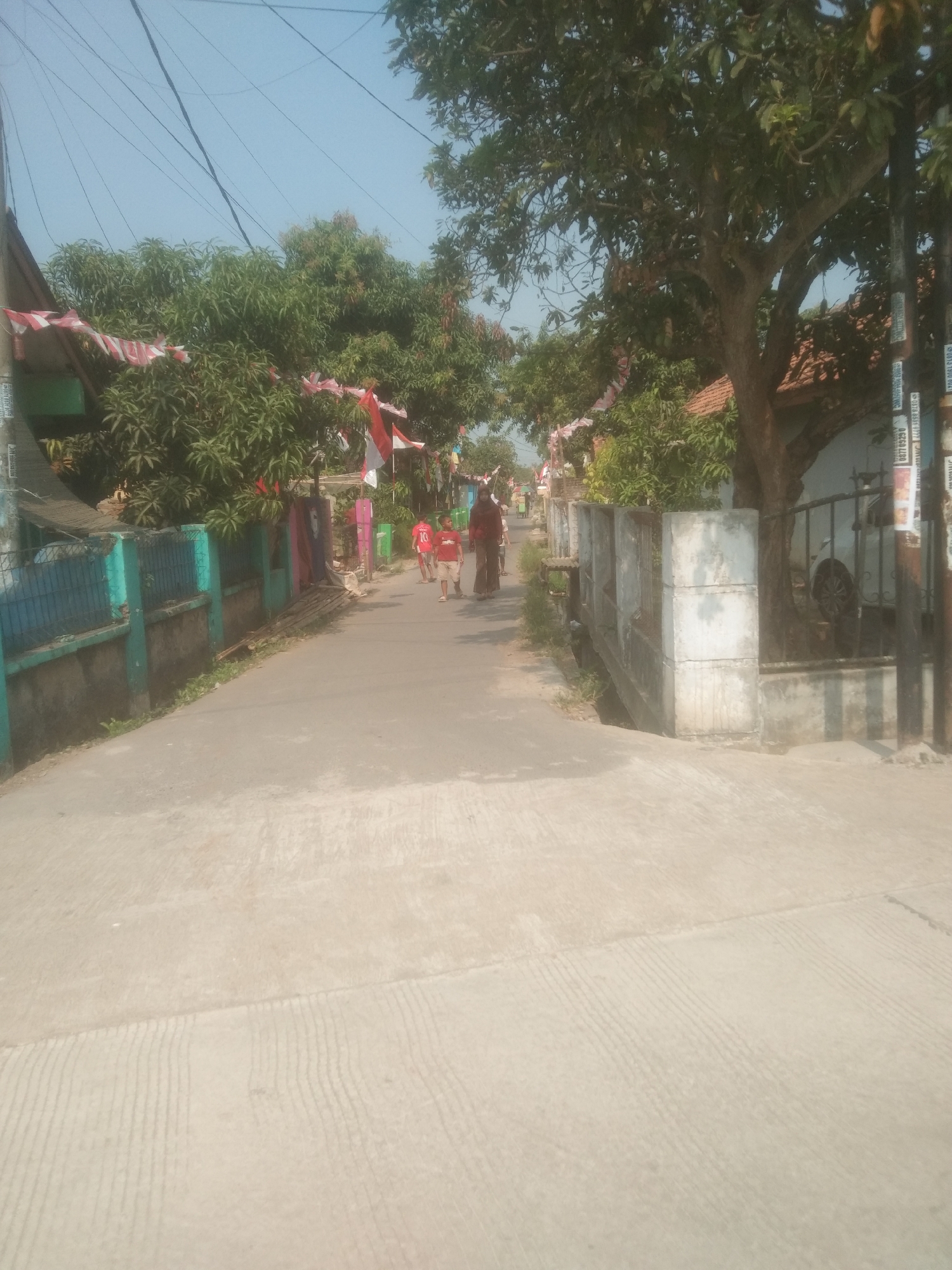 The image depicts a sunlit, paved street in a residential area, likely in Subang, Indonesia, given the presence of Indonesian flags as decorations. The street is lined with houses and lush trees. In the foreground, the concrete pavement shows textured lines, possibly for traction or drainage.
Several people are visible walking along the street. Two young individuals, dressed in red shirts (one with the number 10), are walking ahead, while an adult in a dark headscarf and dark pants walks behind them. Further down the street, other figures are present, and more flags are visible, suggesting a festive occasion or national celebration. The houses have traditional tile roofs. A turquoise fence runs along the left side of the street, bordering a property with dense green foliage. On the right side of the street, a concrete wall with square pillars and a metal gate partially obstructs the view of a house and a car parked behind it. The sky is clear and bright blue.