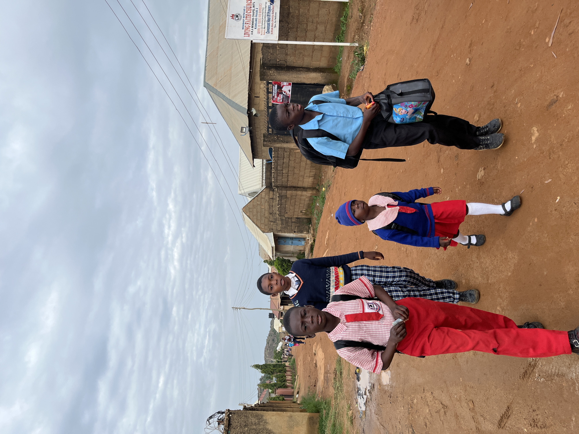 In Jos, Nigeria, four children, three boys and one girl, walk along a dirt road. The two closest children are boys wearing school uniforms. The boy on the left is wearing a red and white checkered shirt, a red tie, and red pants, with a black backpack. The boy to his right is wearing a navy blue sweater with a white collar and a patterned band, and checkered pants. Behind them, a younger boy in a blue sweater, blue hat, and red shorts walks alongside a younger girl also in a school uniform. The girl is wearing a white shirt with a red and white checkered collar, a blue sweater, and a red skirt, with white socks and black shoes. The boy on the far right, also in a school uniform, wears a light blue collared shirt and black pants, carrying a black backpack with a cartoon character attached. The background shows a residential area with brick houses, corrugated metal roofs, and a sign for "LIVING FAITH CHURCH". The road is unpaved and muddy in places, with some scattered debris. The sky is overcast with gray clouds.