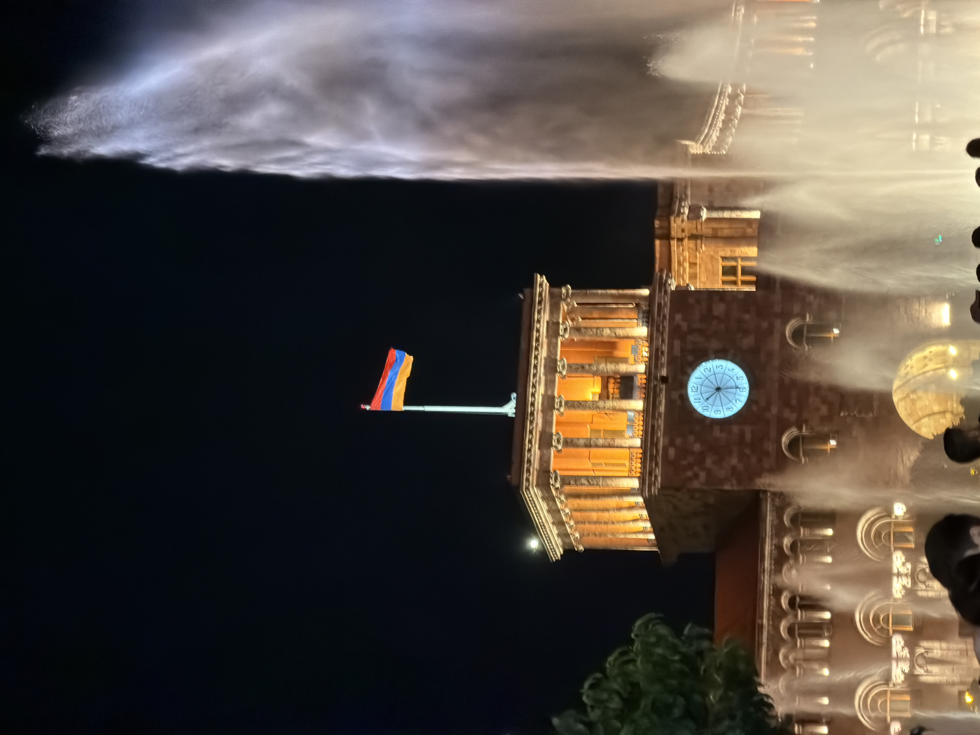 The image captures a vibrant nighttime scene in Yerevan, Armenia, showcasing a grand illuminated building and a spectacular water fountain display. The central subject is a large, stately building, possibly the Government House in Republic Square, which occupies the right side of the frame. It is bathed in warm golden light, highlighting its classical architectural features, including numerous columns, arched windows, and a prominent clock tower. A round clock face on the building displays numbers from 1 to 12, though the exact time is indistinct.
To the left of the building, a powerful water fountain sends a massive jet of water high into the dark night sky. This voluminous spray is brightly illuminated, creating a glowing, misty cloud that stands out against the black background and partially obscures other parts of the building.
A significant national symbol, the Armenian flag, with its distinctive red, blue, and orange horizontal stripes, proudly flies from a flagpole atop the illuminated building. In the bottom right foreground, dark shapes of trees or bushes are visible, along with indistinct dark forms that appear to be the heads or shoulders of people, suggesting spectators enjoying the event.
The scene depicts a lively public event, likely a "singing fountains" show, a well-known attraction in Yerevan's Republic Square. The atmosphere is one of grandeur and national pride, perfectly encapsulated under the clear night sky.