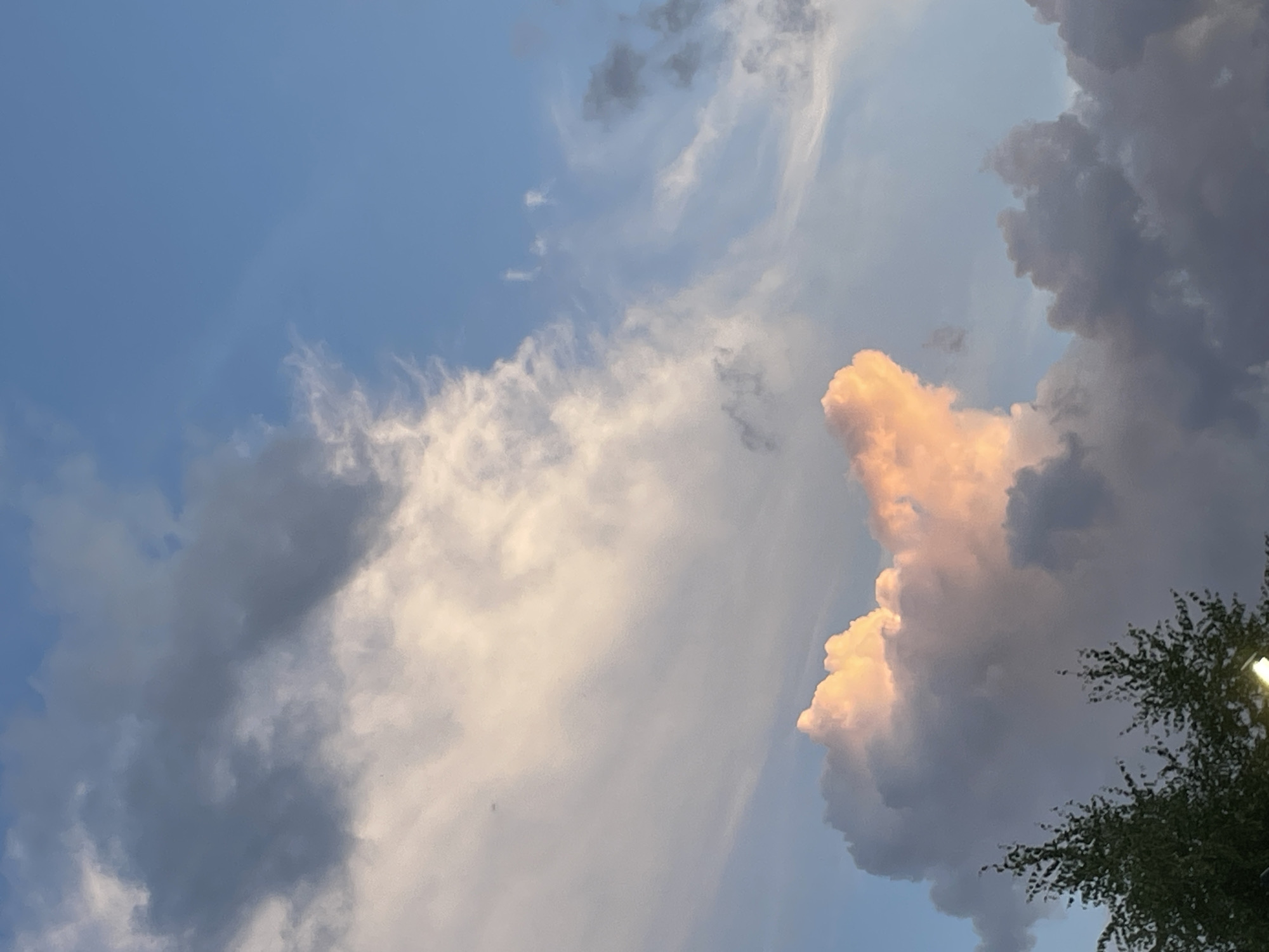 The image shows a partly cloudy sky with a few trees visible in the foreground. The clouds are illuminated by the setting sun, with hues of pink and orange blending into the blue sky. The location is the city of Laishevo, Russia.