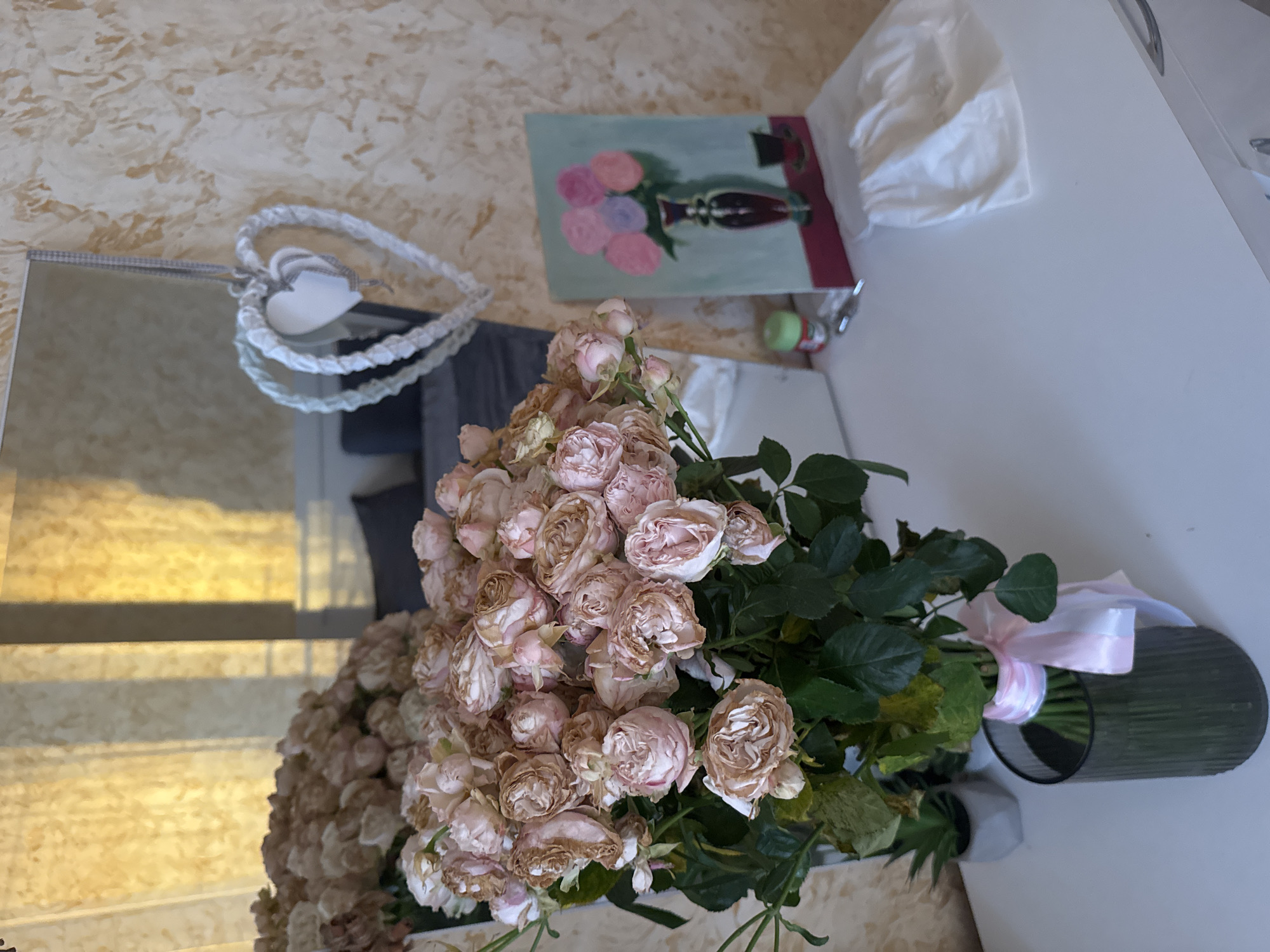 The image shows a bouquet of faded roses in a dark-colored vase with a light pink ribbon tied around it, placed on a white surface. To the left is a mirror reflecting a decorative heart and a glimpse of a room with a patterned wall. Further in the background is a painting of a vase with flowers and a crumpled white cloth. The location is Unknown, Unknown.