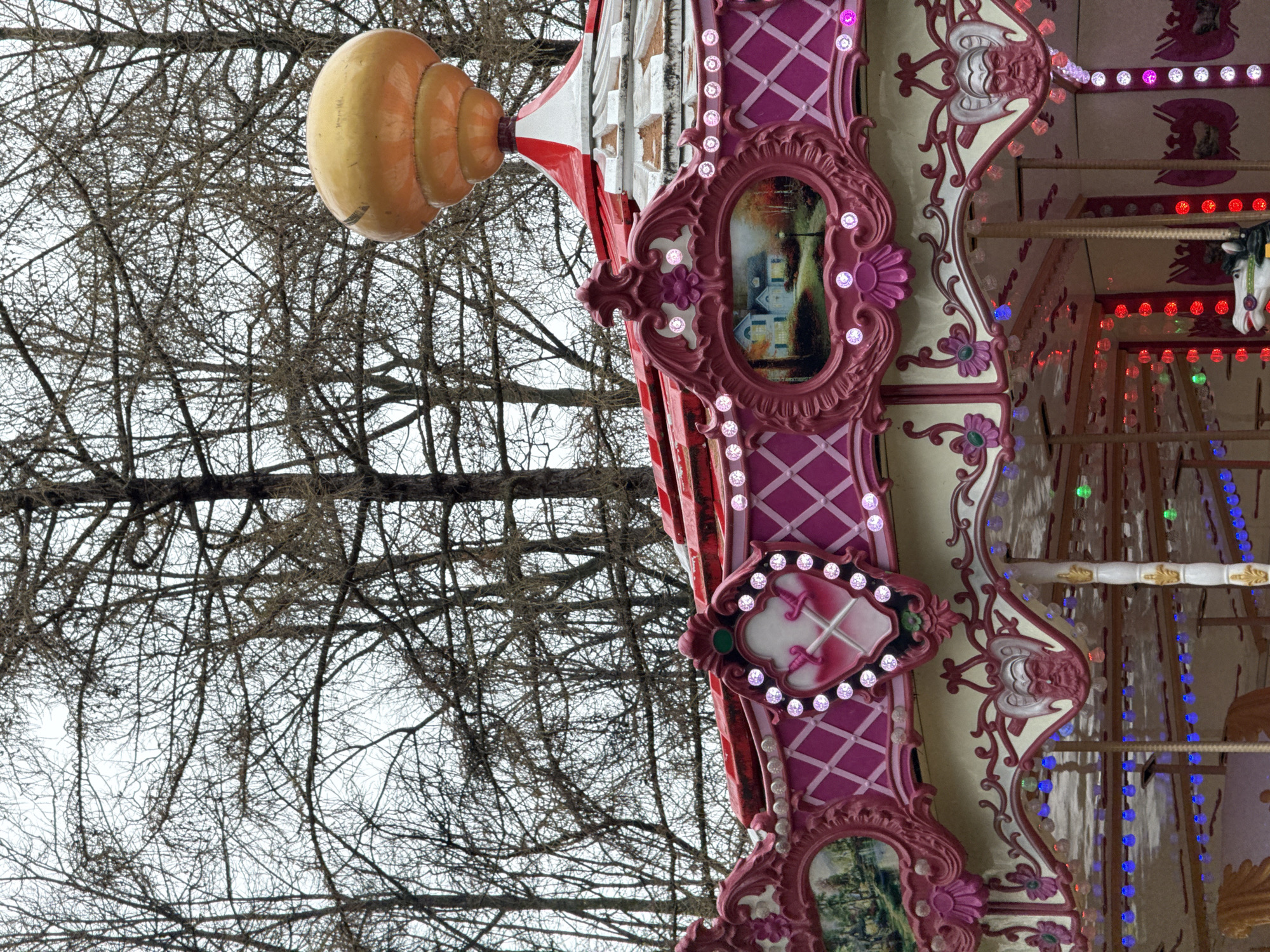 The image captures a section of a merry-go-round located in Perm, Russia, with bare tree branches in the background. The merry-go-round is decorated in a whimsical style, with a color scheme of pink, white, red, and tan. Elaborate detailing is evident, including ornate frames, lattice patterns, and embedded lights. The scene appears to be set outdoors, with the bare trees suggesting either late autumn or winter. The image is taken at an upward angle. A large, layered, tan-colored, onion-like structure is visible at the top left.