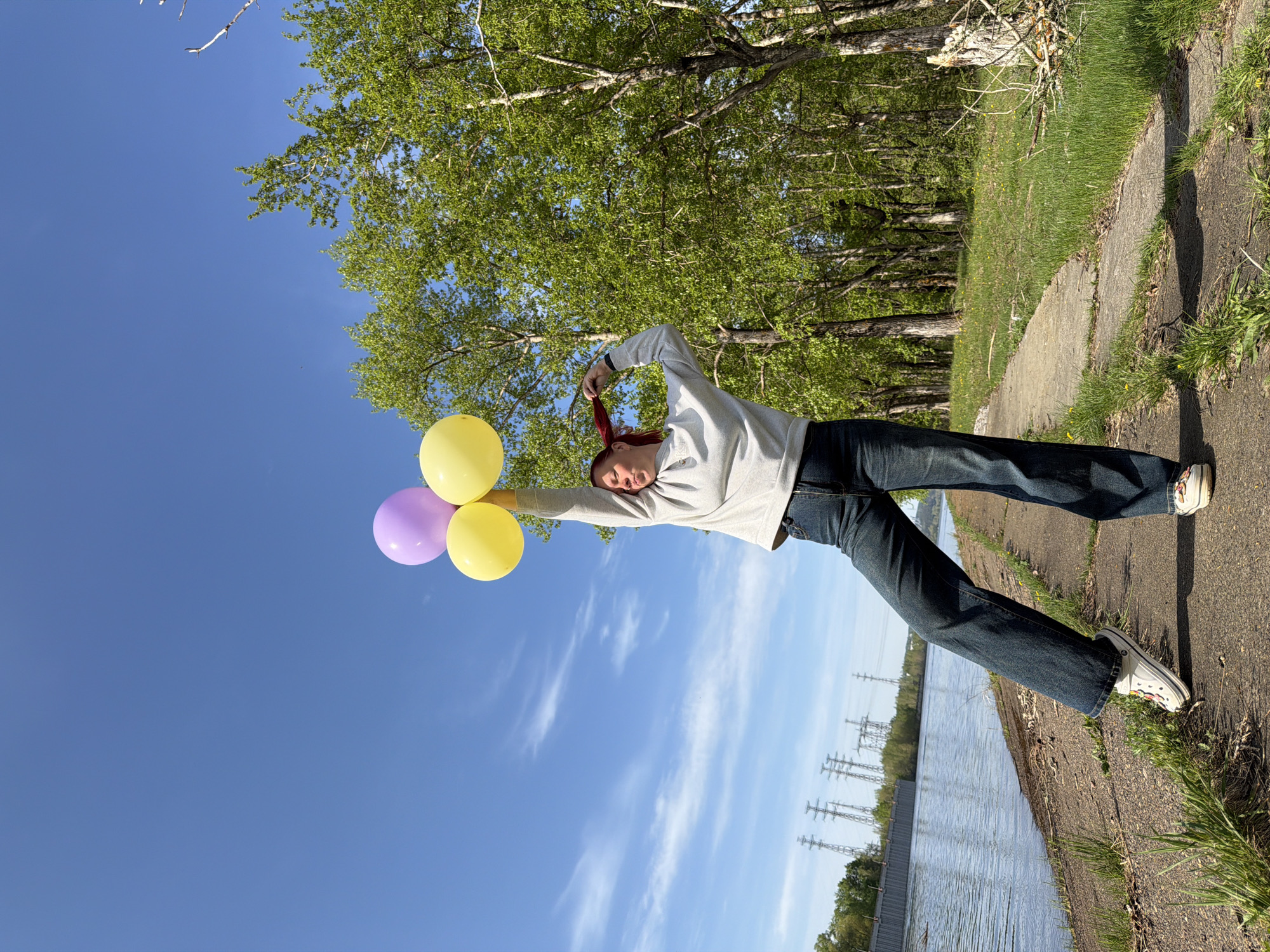 A woman stands on a concrete path near a body of water in Chaikovsky, Russia. She holds a bunch of balloons in her left hand, comprising two yellow and one lavender balloon. She pulls on her red hair with her right hand. The woman wears a gray sweatshirt and blue jeans, with white Crocs on her feet. A line of trees and power lines runs along the opposite side of the water. The sky is blue with some clouds.
