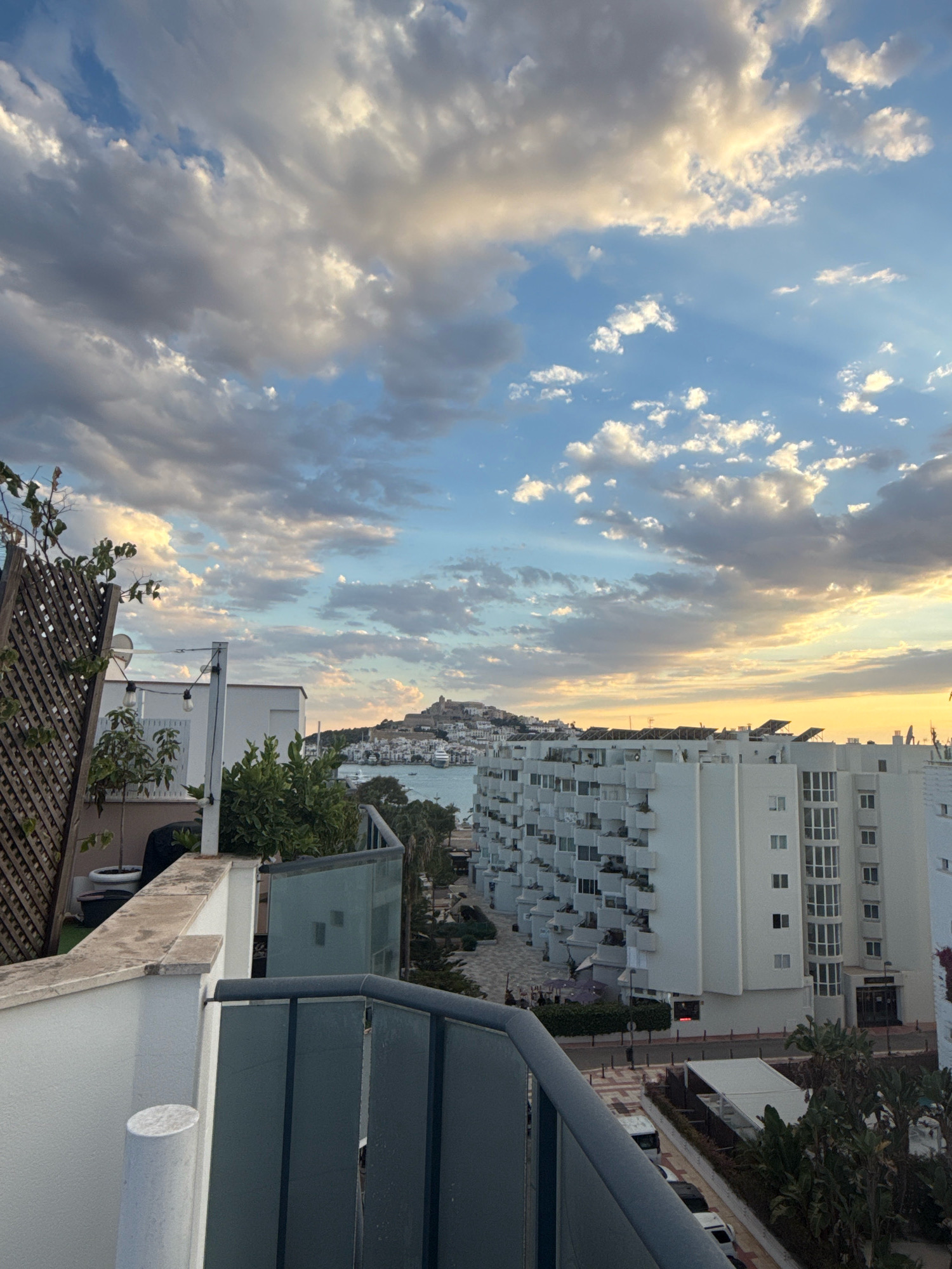 This is a high-angle, eye-level, outdoor, medium shot of a residential building in Ibiza, Spain, likely taken from a balcony or rooftop. The sky is filled with dramatic clouds, with streaks of sunlight breaking through, suggesting either dawn or dusk. In the distance, across a body of water, a fortified hill town is visible. In the foreground and mid-ground are modern white apartment buildings with numerous balconies. Some vegetation is present on the balconies and in the foreground. A dark-railed balcony railing runs across the bottom of the image. The lighting suggests it is late afternoon or early evening.
