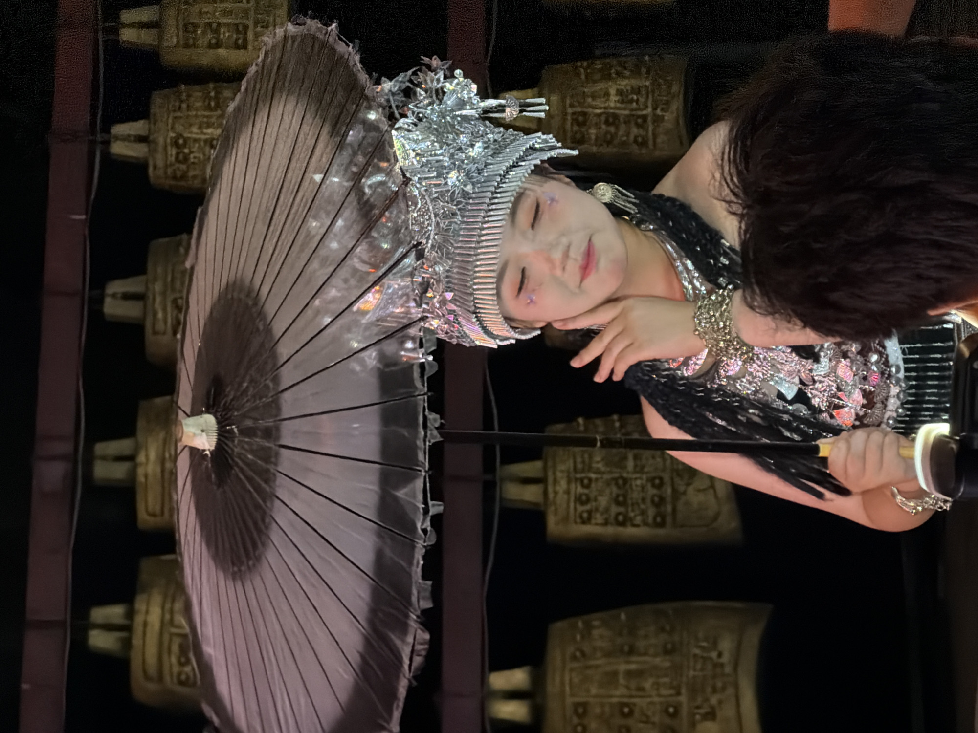 In the city of Yuzhong District, China, a young woman adorned in traditional attire, including an ornate silver headdress and sparkling costume, holds a translucent umbrella over her head. She is positioned in front of a display of numerous large, ornate bronze bells, suggesting a cultural performance or exhibition. The woman's pose, with her hand gently touching her face, conveys a sense of grace and poise. The background, with the dimly lit bells, creates an atmosphere of historical significance and tradition.