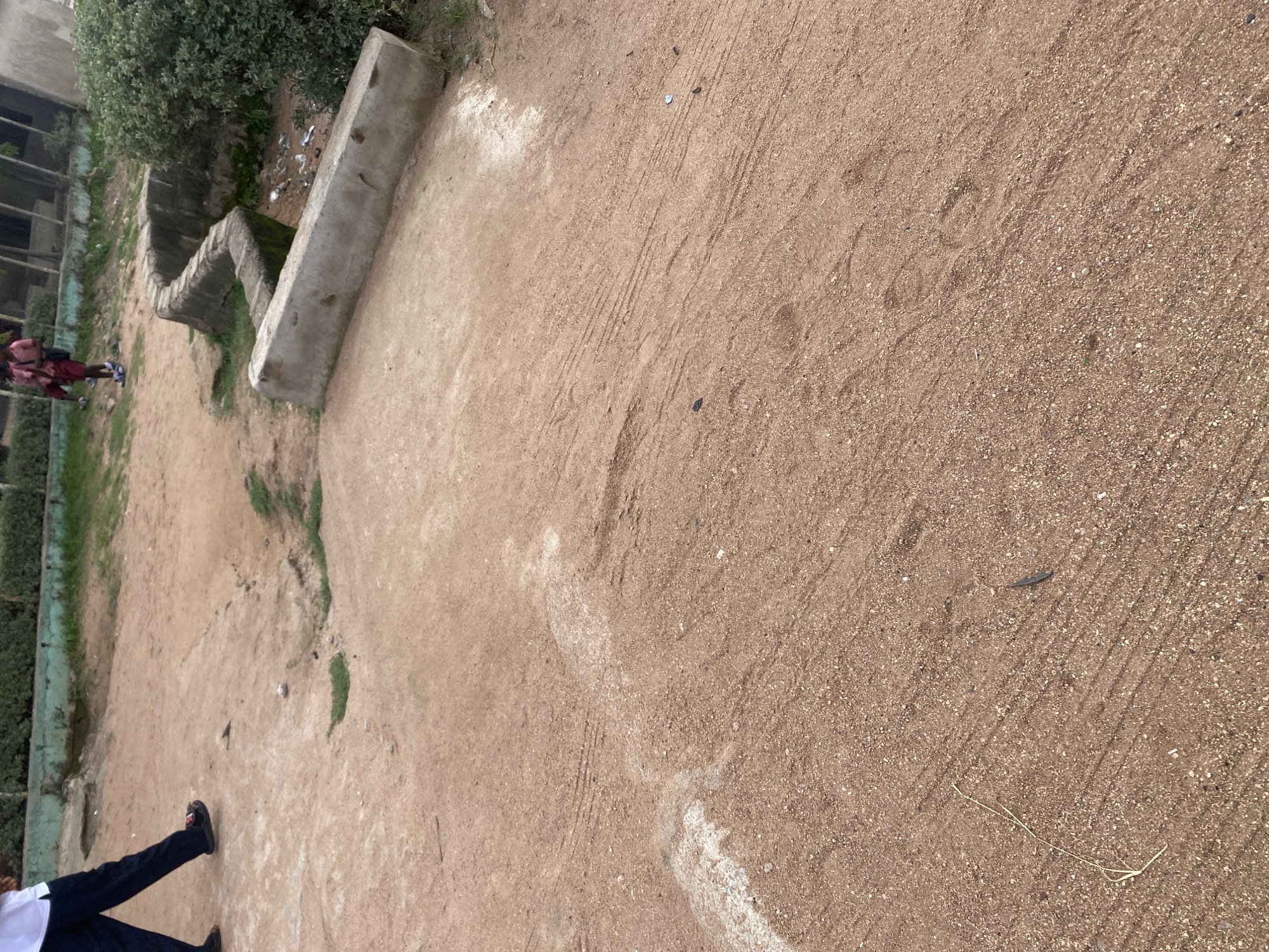 The image depicts an outdoor scene in Jauro Yinu, Nigeria, characterized by a sandy ground surface with visible tire tracks and some small pebbles and debris scattered across it. In the background, a zig-zagging concrete structure, possibly a planter or a part of an urban design, is visible. Adjacent to this structure is a patch of green vegetation, suggesting a somewhat unkempt outdoor area. Further in the background, parts of a building with a faded green border are discernible.
In the foreground and mid-ground, there are two individuals. One person, wearing dark pants and black shoes, is walking towards the left side of the frame. Another person, dressed in a red and patterned outfit and carrying a backpack, is also present, appearing to walk or stand near the concrete structure. The presence of people suggests a public or communal space.
The lighting suggests it is daytime, with the sun casting shadows. The overall impression is of a casual, everyday moment in a rural or semi-urban setting in Nigeria. No specific event is clearly indicated, and the weather appears to be clear. There is no visible text in the image. The scene is notable for its natural ground texture and the utilitarian concrete element.