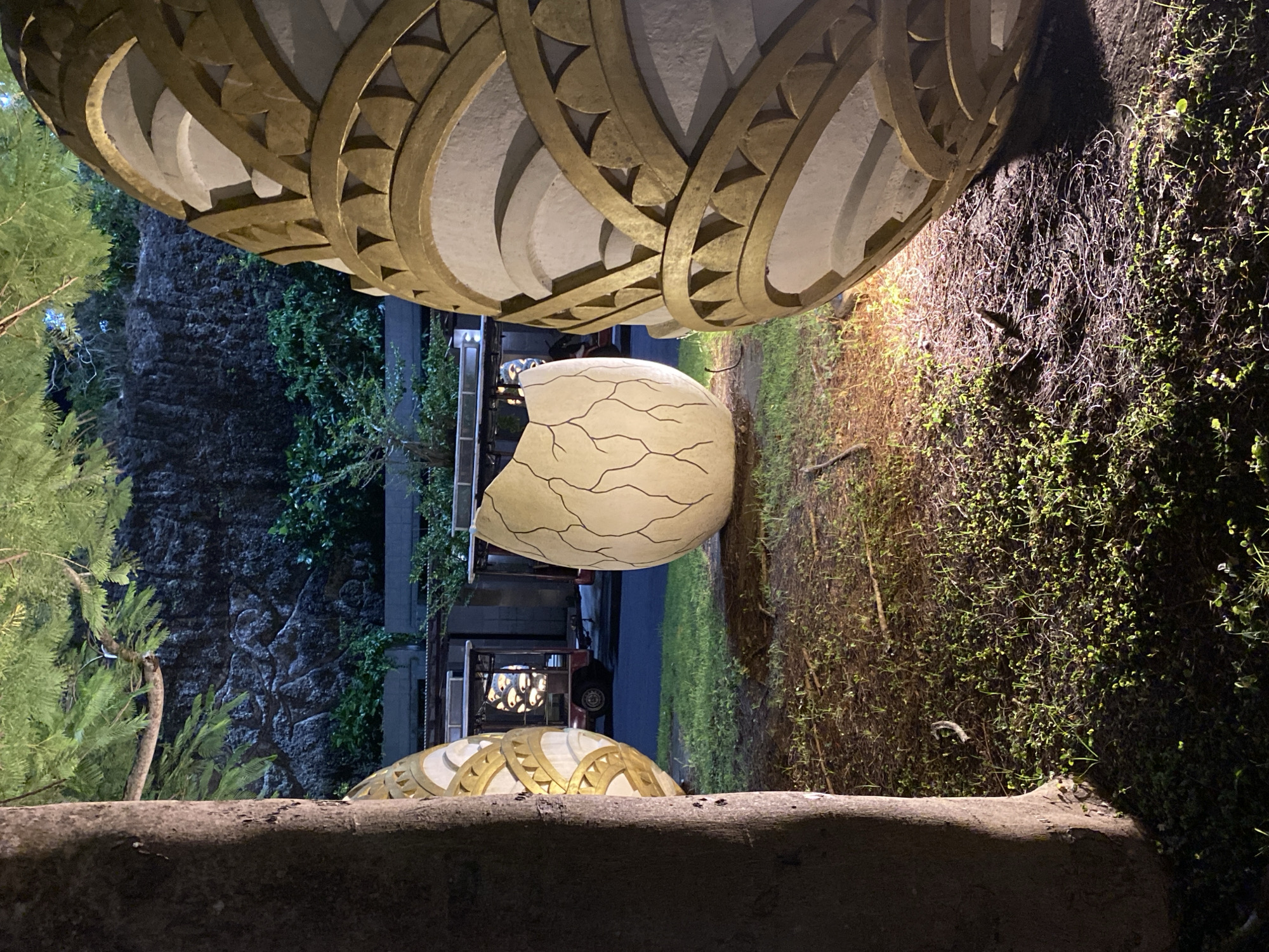 This media file, taken in Kuta Selatan, Indonesia, captures an outdoor scene at night. The primary subjects are two large, decorative egg-shaped objects, illuminated by artificial light. One egg, cracked open, reveals a detailed, light-colored interior with intricate line patterns resembling veins or cracks. The other egg, partially visible at the bottom of the frame, features a similar decorative pattern of golden, crescent-shaped elements against a white background.
The objects are situated in a garden-like setting. To the left, lush green foliage and trees are visible, partially obscuring a dark, textured wall that appears to be made of rock. To the right, the ground is covered with dense, low-lying vegetation, some of which is illuminated by a warm, yellow light source casting shadows. In the background, a dark road is visible, with a vehicle, possibly a cart or small truck, parked on it, also illuminated by internal lights.
The overall mood is somewhat mysterious and serene, enhanced by the nighttime setting and focused lighting. The prominent decorative elements suggest an artistic installation or a festival display. There are no people visible in the frame, and the weather appears to be calm, given the clear visibility and lack of rain. The location cues, such as the tropical-looking foliage, combined with the provided context, place the scene in Kuta Selatan.