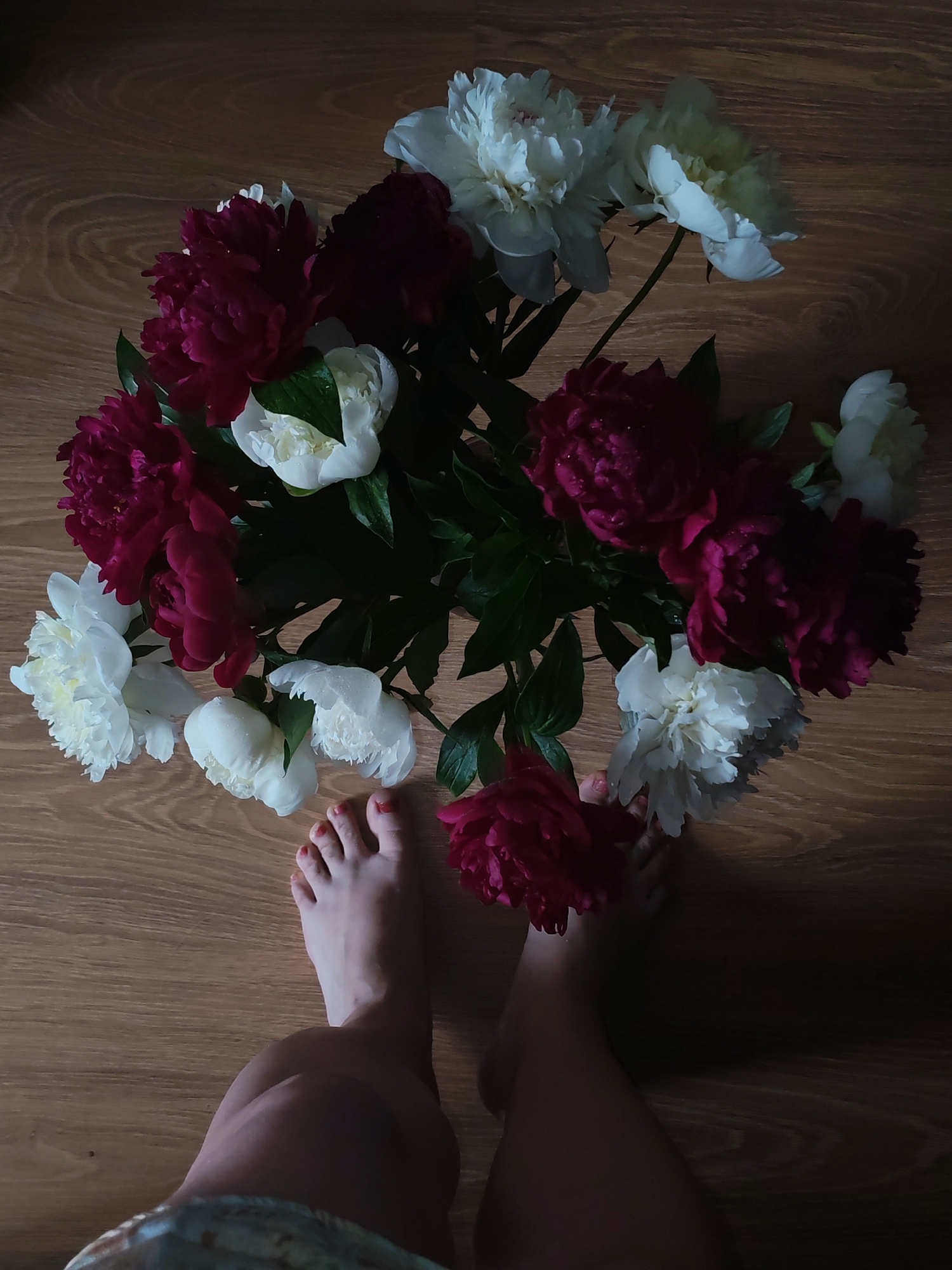 In Izhevsk, Russia, an overhead shot shows a bouquet of peonies, both white and deep pink, arranged on a wooden floor. The flowers are lush and some appear to have water droplets on their petals. In the foreground, a person's bare feet, with red nail polish on the toes, are visible, along with a portion of their legs and a hint of patterned fabric. The lighting is dim, casting shadows and creating a moody atmosphere.