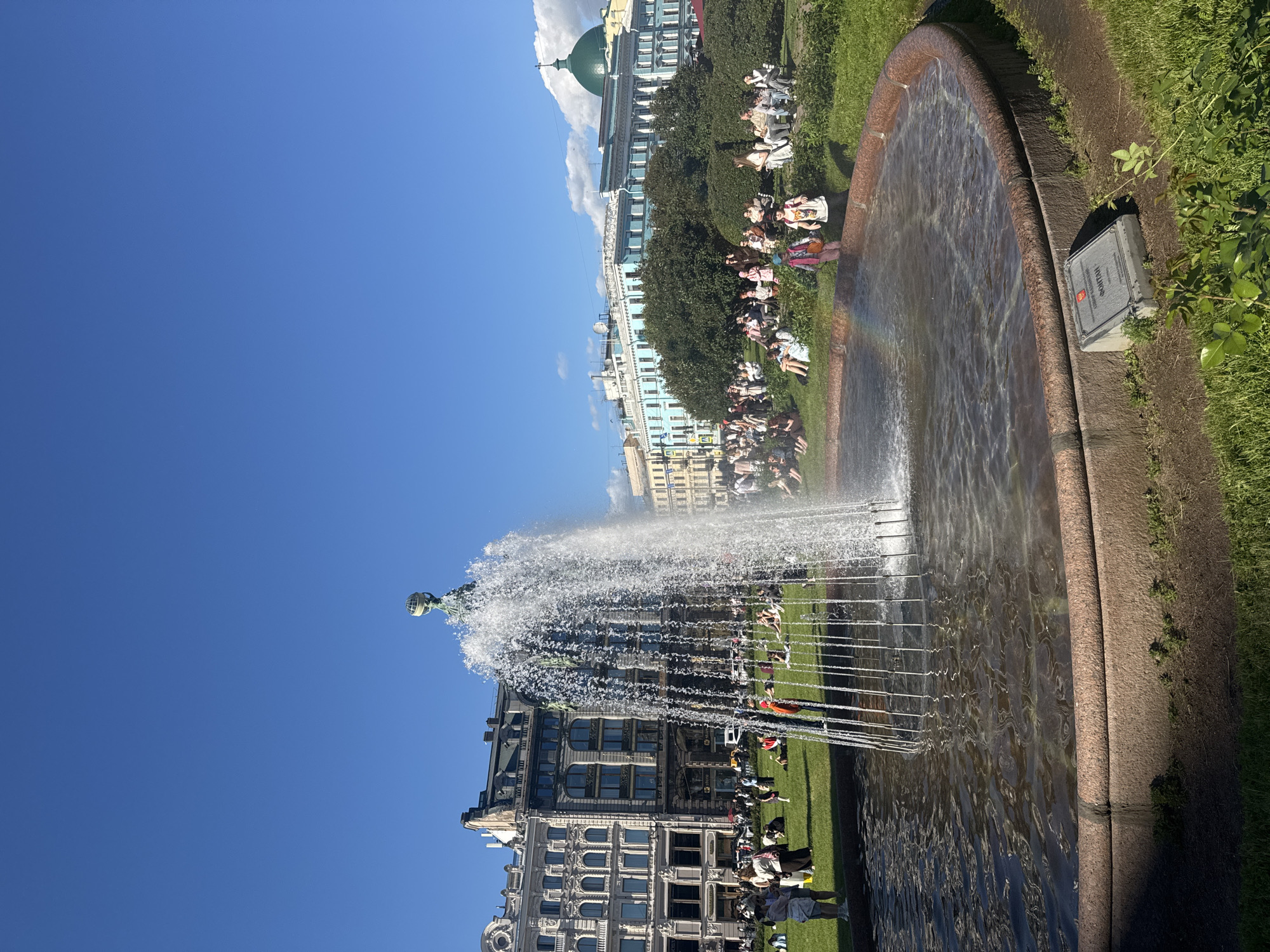 The image depicts a large, tiered fountain with water spraying upwards in a public square on a sunny day. The fountain is situated in front of several multi-story buildings with neoclassical architecture, characteristic of Saint Petersburg, Russia. In the background, a domed building with a green roof is visible, possibly a church or governmental building. People are seen scattered across a grassy area surrounding the fountain, some sitting, some standing, and a few appear to be picnicking. The sky is a clear blue with a few scattered clouds. A faint rainbow is visible within the spray of the fountain. A plaque with text and an emblem is present in the foreground, near the edge of the fountain basin. The overall scene suggests a leisurely afternoon in a historical urban setting.