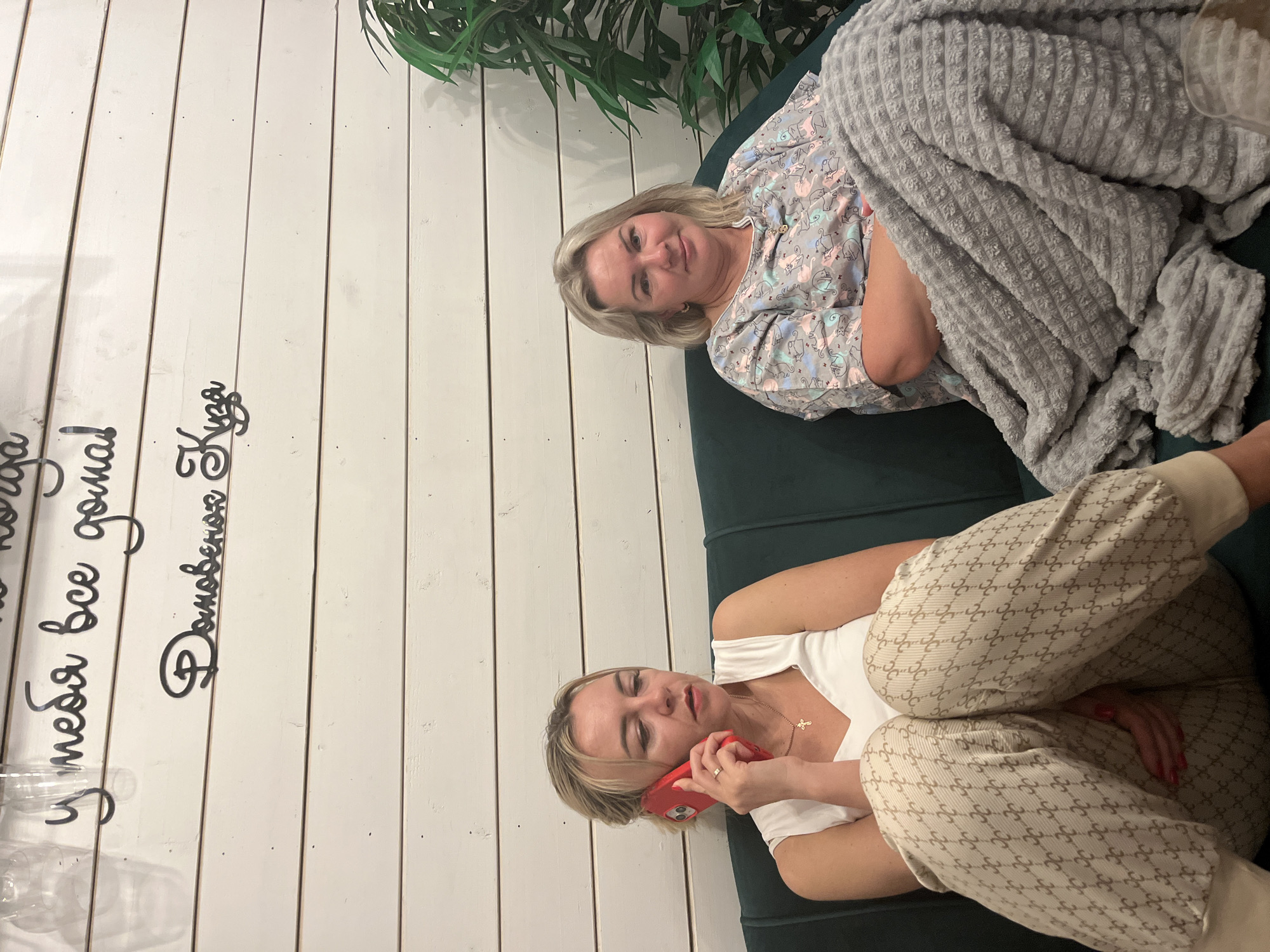 An interior scene in Kamyennoye, Russia, features two adult women seated on a dark green upholstered sofa against a white, horizontally paneled wall.
On the left, a woman with blonde hair, estimated to be in her 30s or 40s, is engaged in a phone call, holding a red smartphone to her right ear. She wears a white sleeveless top and light-colored sweatpants adorned with a repeating pattern resembling interlocking 'G's or similar monograms. A gold necklace with a small cross pendant is visible around her neck. Her expression is focused or serious.
To her right, a second woman, with blonde, shoulder-length hair and appearing to be in her late 40s or 50s, is seated, looking towards the viewer with a slight smile. She wears a light-colored short-sleeved top patterned with various pastel-colored cats (pink, blue, grey). Her lower body is covered by a grey, textured, plush blanket, under which her arms are crossed.
The white paneled wall behind them features black script-style wall decals. The upper visible text reads "у тебя все дома!" (u tebya vse doma!, translating to "Is everything okay at home?" or "Are you all there?"). Below this, the text "Домовенок Кузя" (Domovenok Kuzya) is visible, referring to a popular Russian cartoon character. A green leafy plant is situated in the background on the far right of the frame. The lighting is even, suggesting an indoor setting.