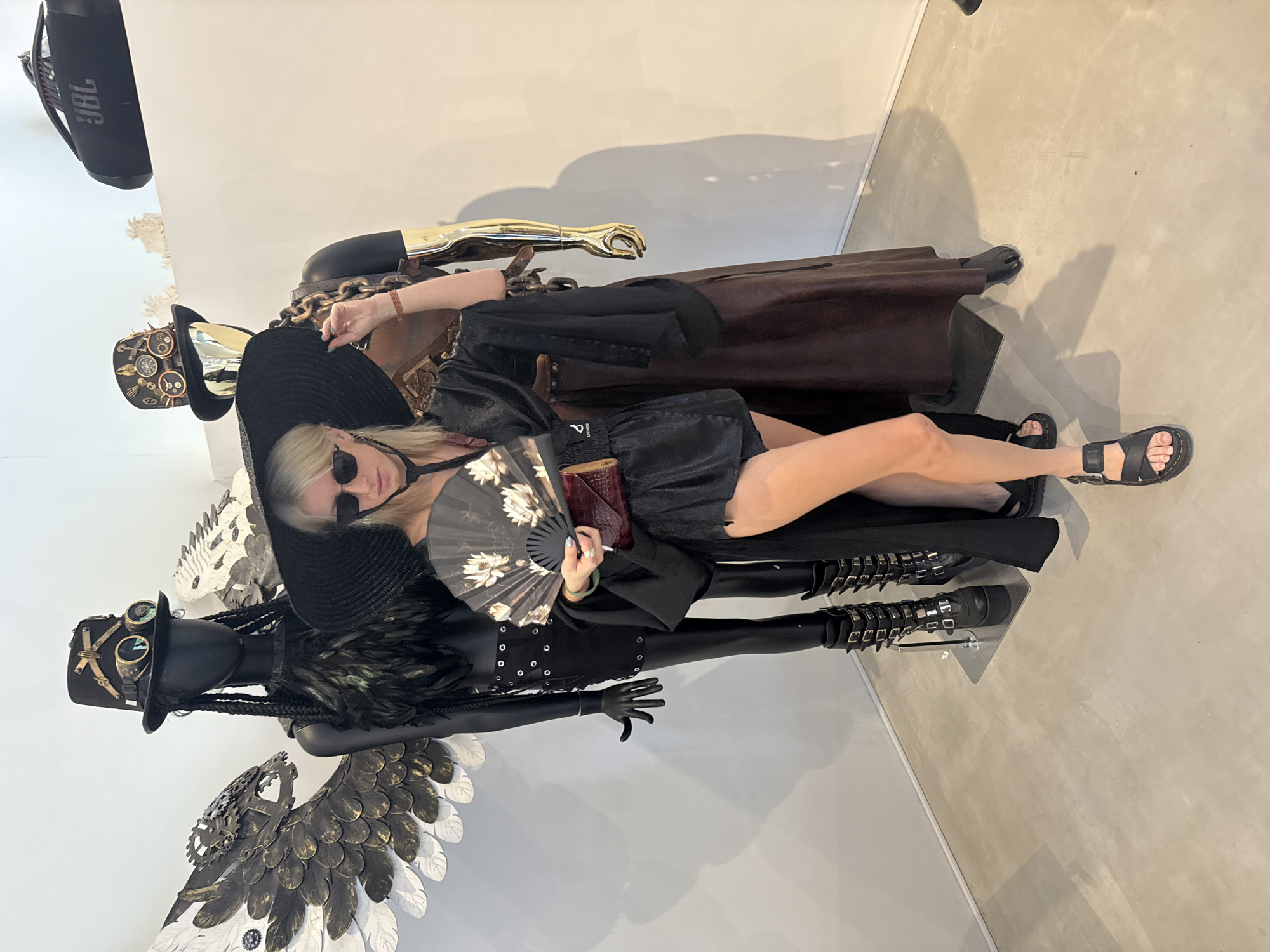 A blonde woman reclines amidst various artistic mannequins or sculptures, posing for a photo indoors, likely in Baan Lipa Noi, Thailand. The image is oriented vertically with the woman lying horizontally across the frame.
The woman wears a large black wide-brimmed hat, black sunglasses, a black top or dress, and black flat sandals. She holds a black fan with white floral patterns in one hand and a small, reddish-brown clutch in the other. Her demeanor appears relaxed and stylish.
To her left, a black mannequin is adorned with a steampunk-inspired hat featuring goggles and gears, alongside large, intricate wings that are a mix of metallic dark feathers and white/silver sections. This mannequin also features long black dreadlocks and a black feathered bodice. Directly behind the woman's head and upper body is another sculptural element, featuring a golden, articulated arm reaching upwards from a dark, cloaked form. Underneath the woman's lower body, and seemingly supporting her, is a dark figure or mannequin leg clad in boots with numerous silver buckles.
The background consists of plain, light-colored walls and a matching light-toned floor. A black JBL speaker is visible hanging from the ceiling in the upper left of the frame. The overall aesthetic is dark, edgy, and artistic, suggesting an exhibition, gallery, or fashion display.