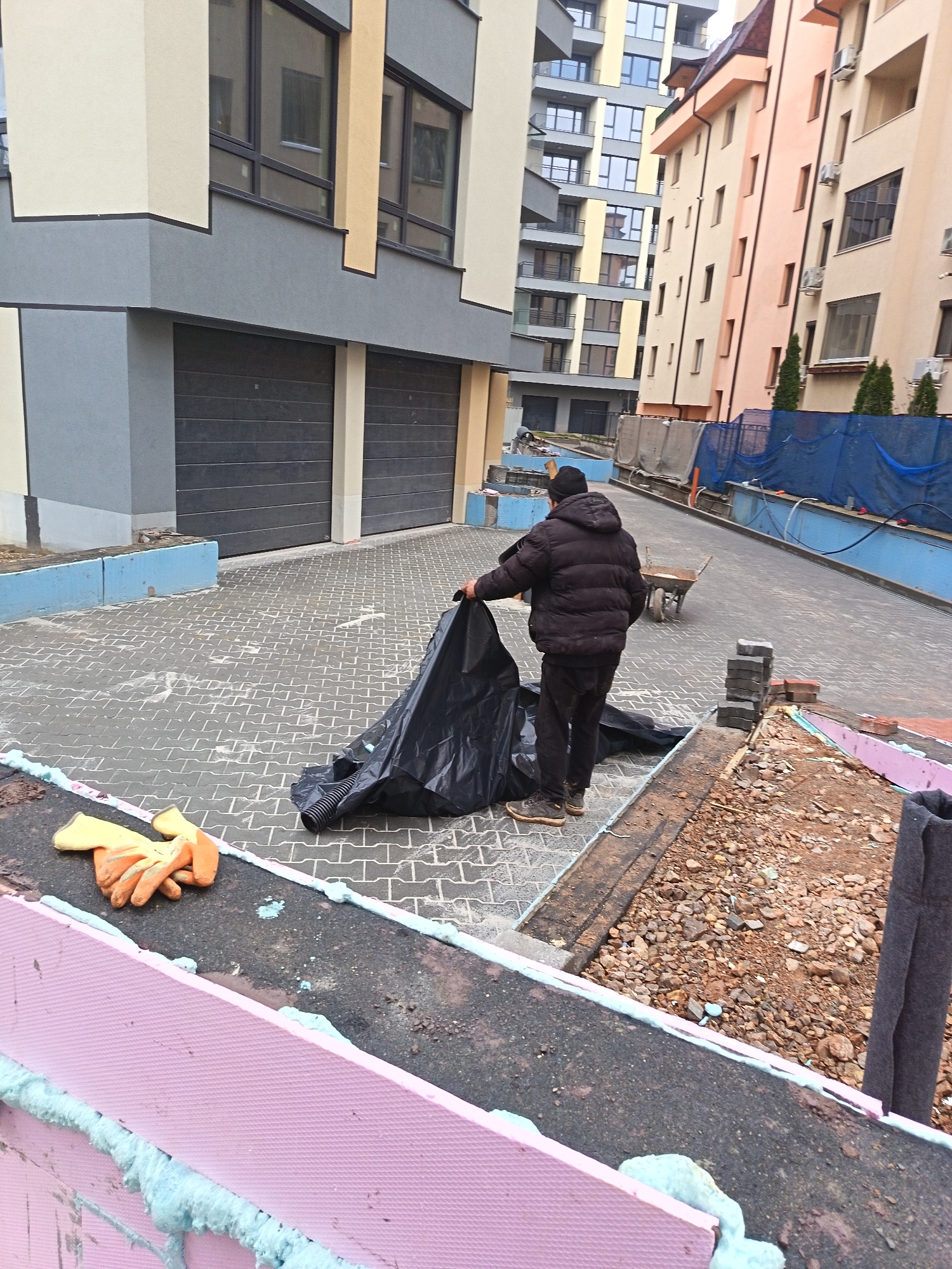 A man in a black puffy jacket and pants is handling a large black plastic sheet on a brick-paved area between buildings. The buildings are multiple stories with modern architecture. The location is likely in Sofia, Bulgaria, based on architectural styles common in the region. In the foreground, there are pink insulation boards with blue foam sealant. Orange work gloves are lying on top of one of the boards. A pile of bricks and construction debris is next to the man. In the background, there is a wheelbarrow and some blue safety netting.