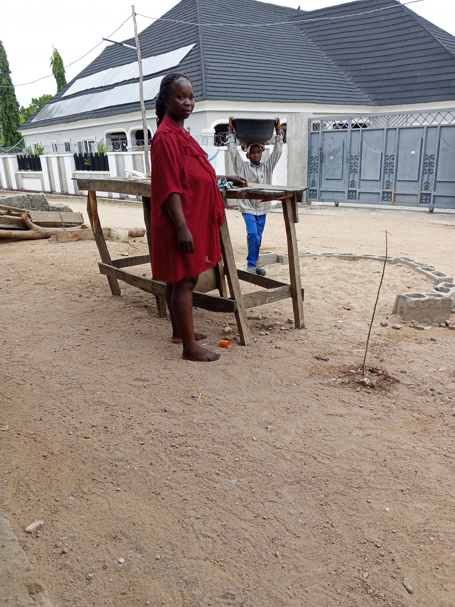 The image depicts a dusty outdoor scene in Adonkolo, Nigeria. A barefoot pregnant woman in a red dress stands to the left of a wooden table. A child stands behind the table, balancing a black tub on their head. A sapling is planted in the sandy ground near a ring of concrete blocks. In the background is a gray gate and a house with solar panels on the roof. Construction debris is scattered on the ground.