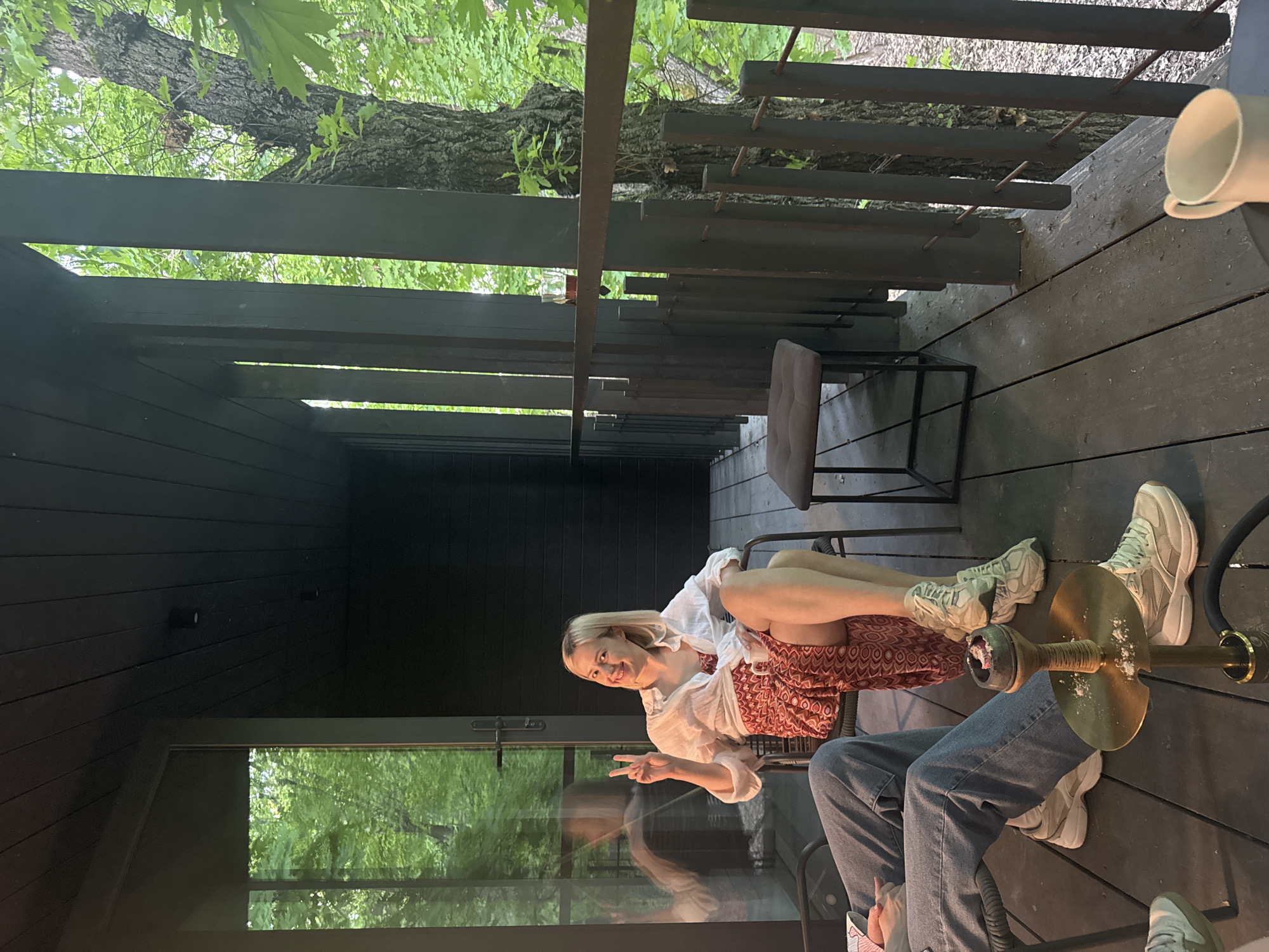 The image captures a scene on a wooden deck, possibly a patio or balcony, potentially located in Усть-Курдюм, Russia. The viewpoint is from above, angled downward towards the scene.
Two people are visible, one is a fair-skinned woman with blonde hair who is smiling and holding up a peace sign. She is wearing a white blouse over a patterned red and brown dress. Next to her, a second person is partially visible, wearing blue jeans.
A hookah sits between the two people on the deck.
The background features dark wooden structures, possibly part of the building or deck, and glimpses of greenery, suggesting trees or foliage nearby. There is also a window visible, reflecting the greenery outside.
A metal chair is visible behind the woman. A cup is on the right.
