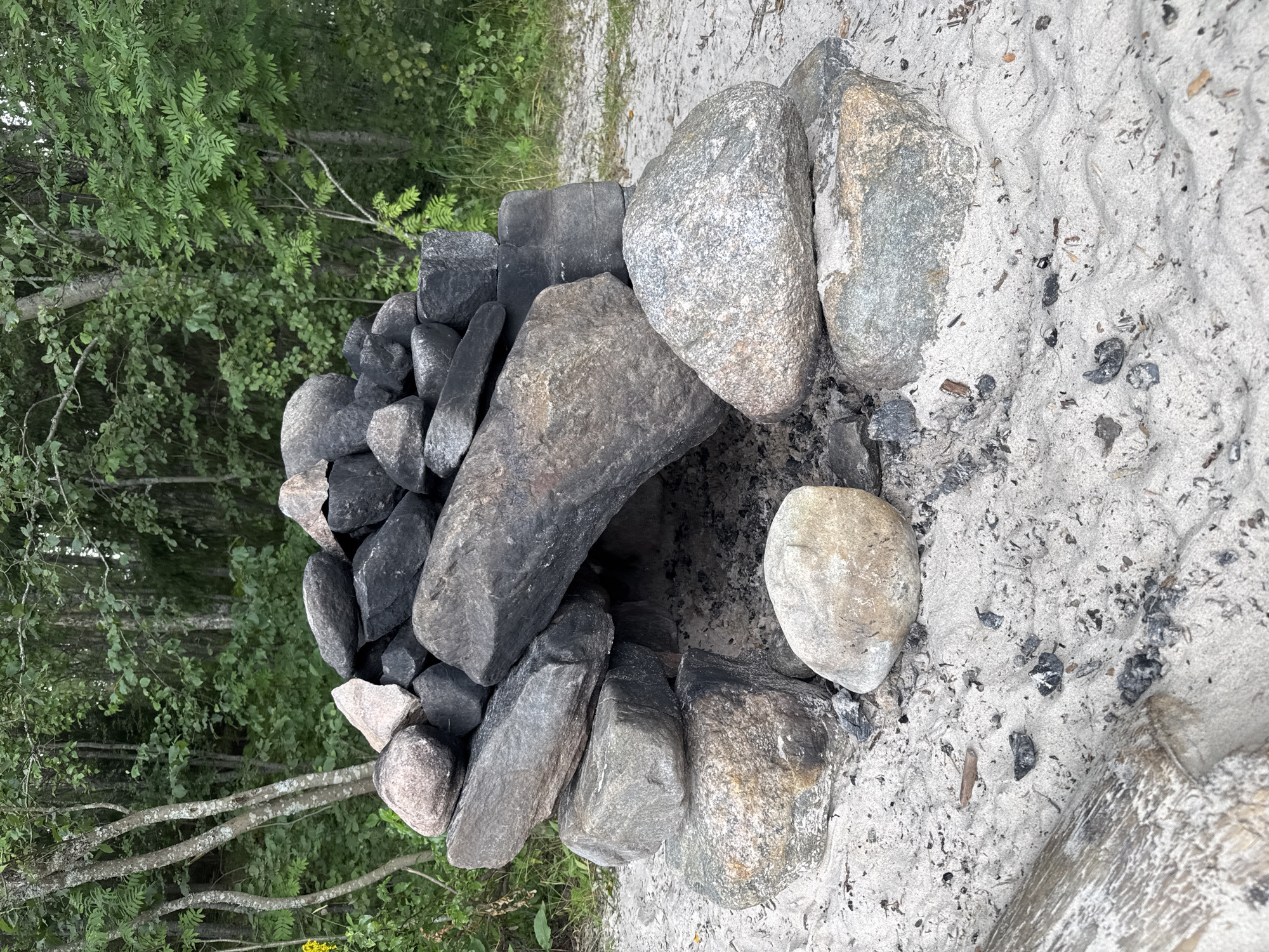 The image shows a fire pit constructed from various large rocks, situated on a sandy area. The rocks are stacked in a rough circular formation with an opening in the center, creating a space for a fire. Some of the rocks appear charred, indicating recent use. The ground surrounding the fire pit is sandy, with scattered pieces of charcoal and debris. In the background, there is a dense forest with green foliage and trees. The location is described as Sorola, Russia, suggesting an outdoor, possibly recreational or wilderness setting. There are no people or animals visible in the image. The overall scene depicts a rustic outdoor fire pit, prepared for or having recently been used for a fire.